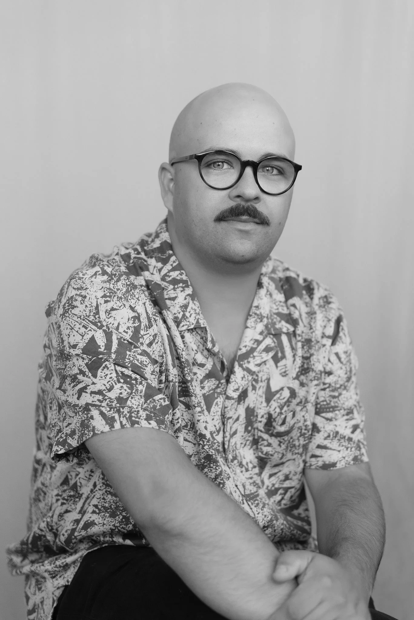Homme chauve avec moustache et lunettes rondes, portant une chemise à motifs graphiques, posant devant un fond clair, portrait du photographe de mariage Vincent Labonté, Photographe mariage Québec.