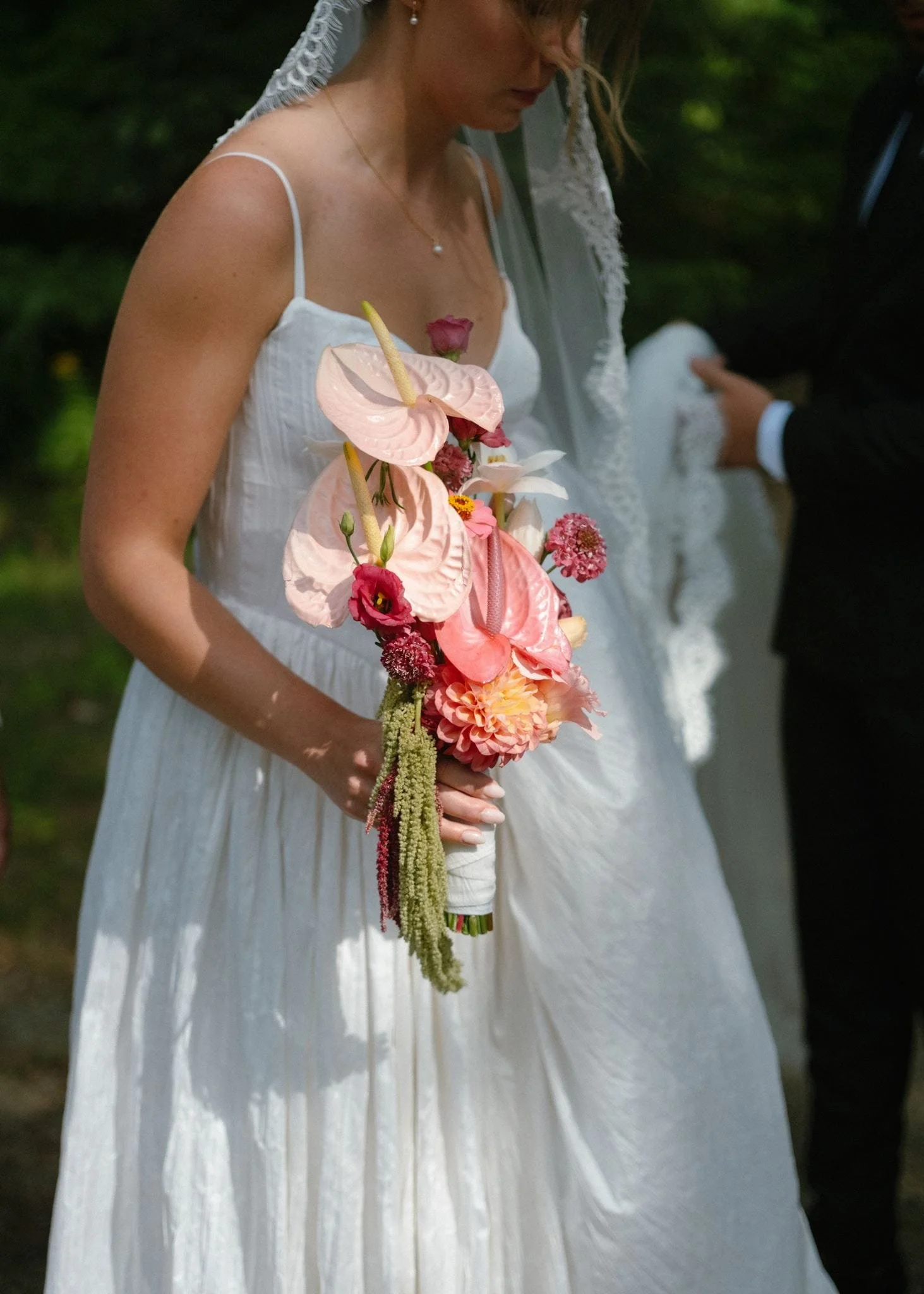 Une femme portant une robe blanche de mariage qui tient un bouquet de fleurs composé de fleurs roses, blanches et rouge foncé dans un environnement vert naturel.