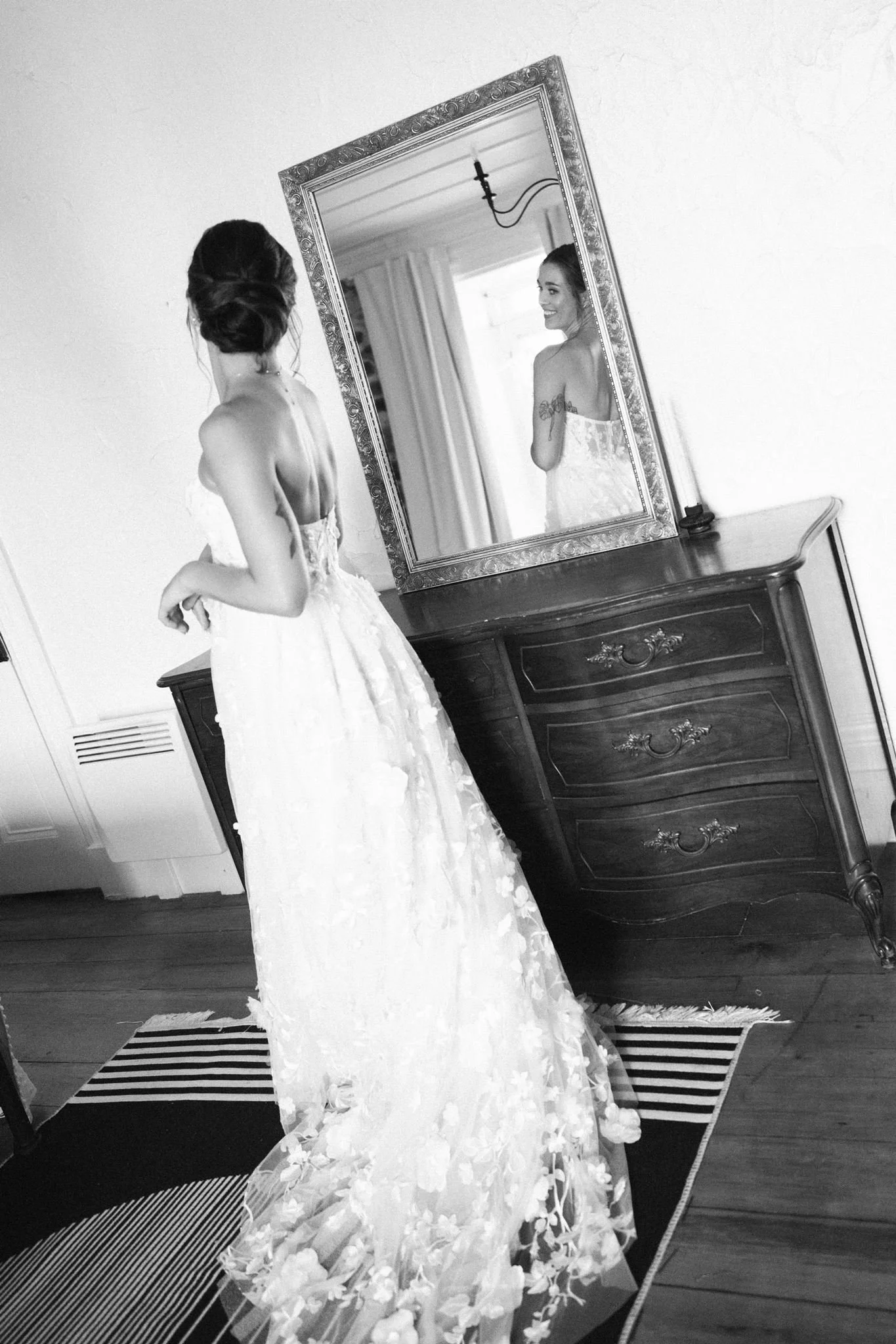 Une femme en robe de mariage regarde dans un miroir, souriant, dans une pièce avec un mobilier en bois foncé.
