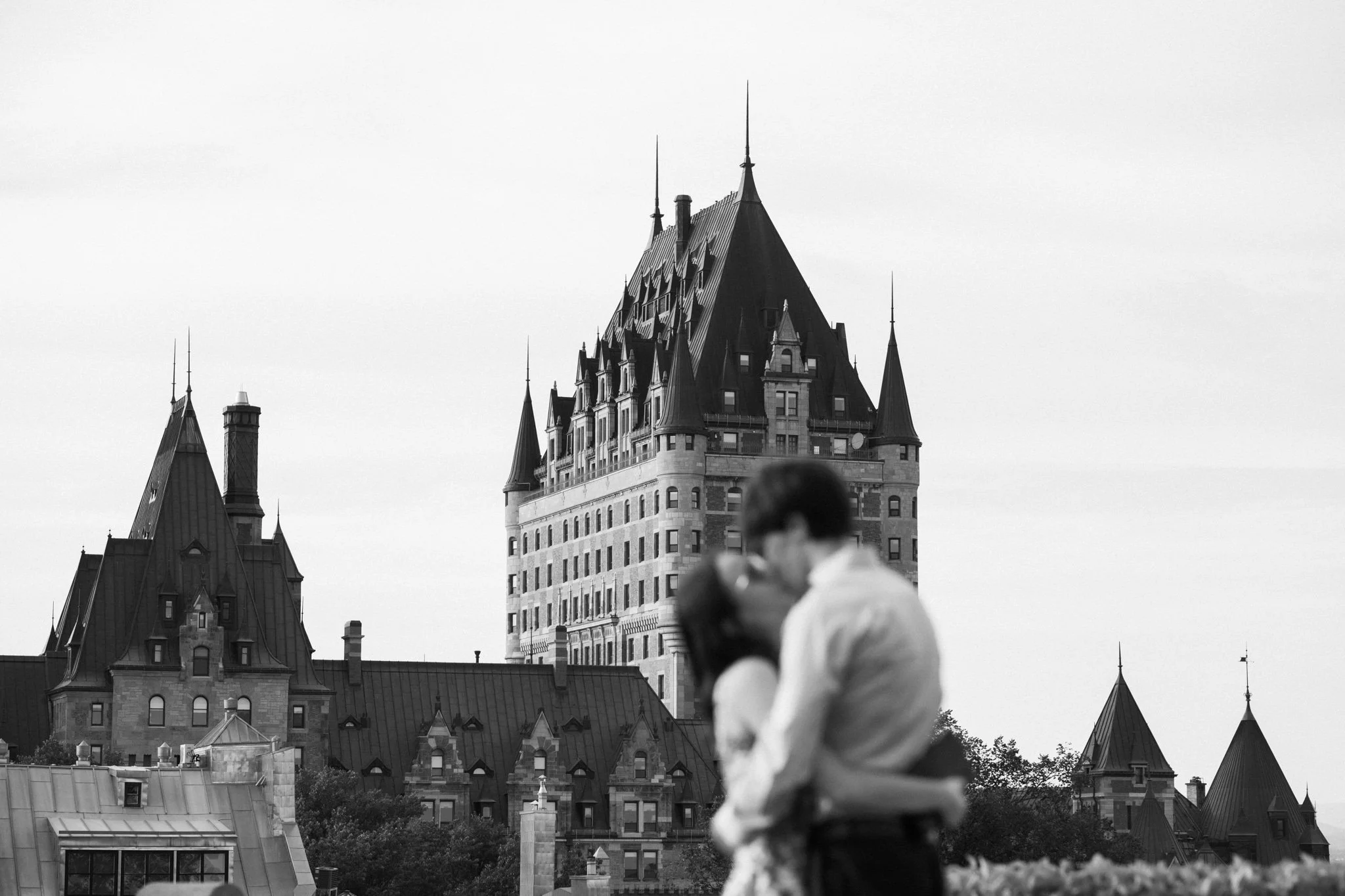 Une photo en noir et blanc d'un château avec des tours pointues et un toit en pente, un homme flou en premier plan, et un ciel clair en arrière-plan.