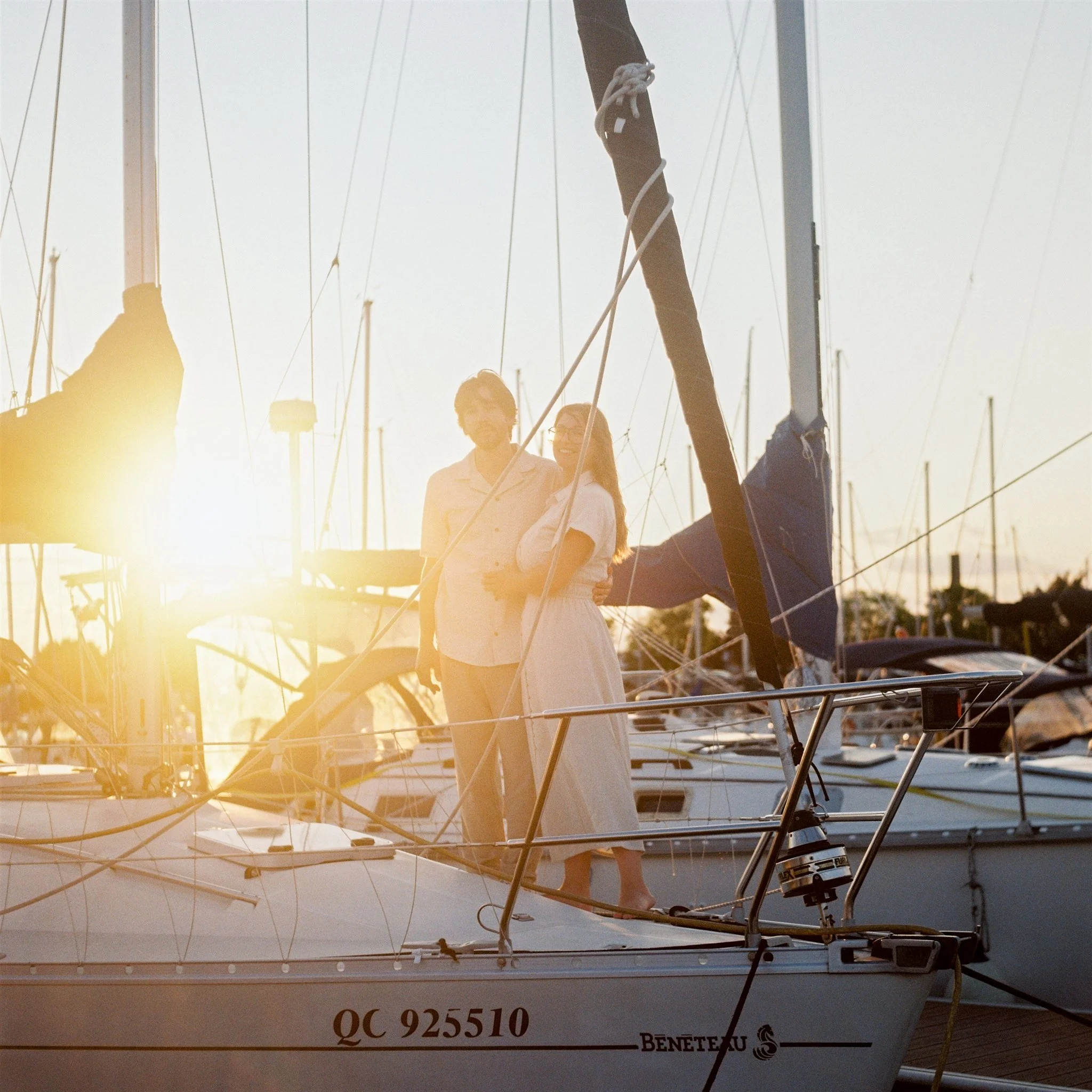 Deux femmes se tiennent à côté d'un bateau à voiles amarré, avec un arrière-plan de bateaux et de lampadaires. Le soleil est bas, créant une lumière dorée.
