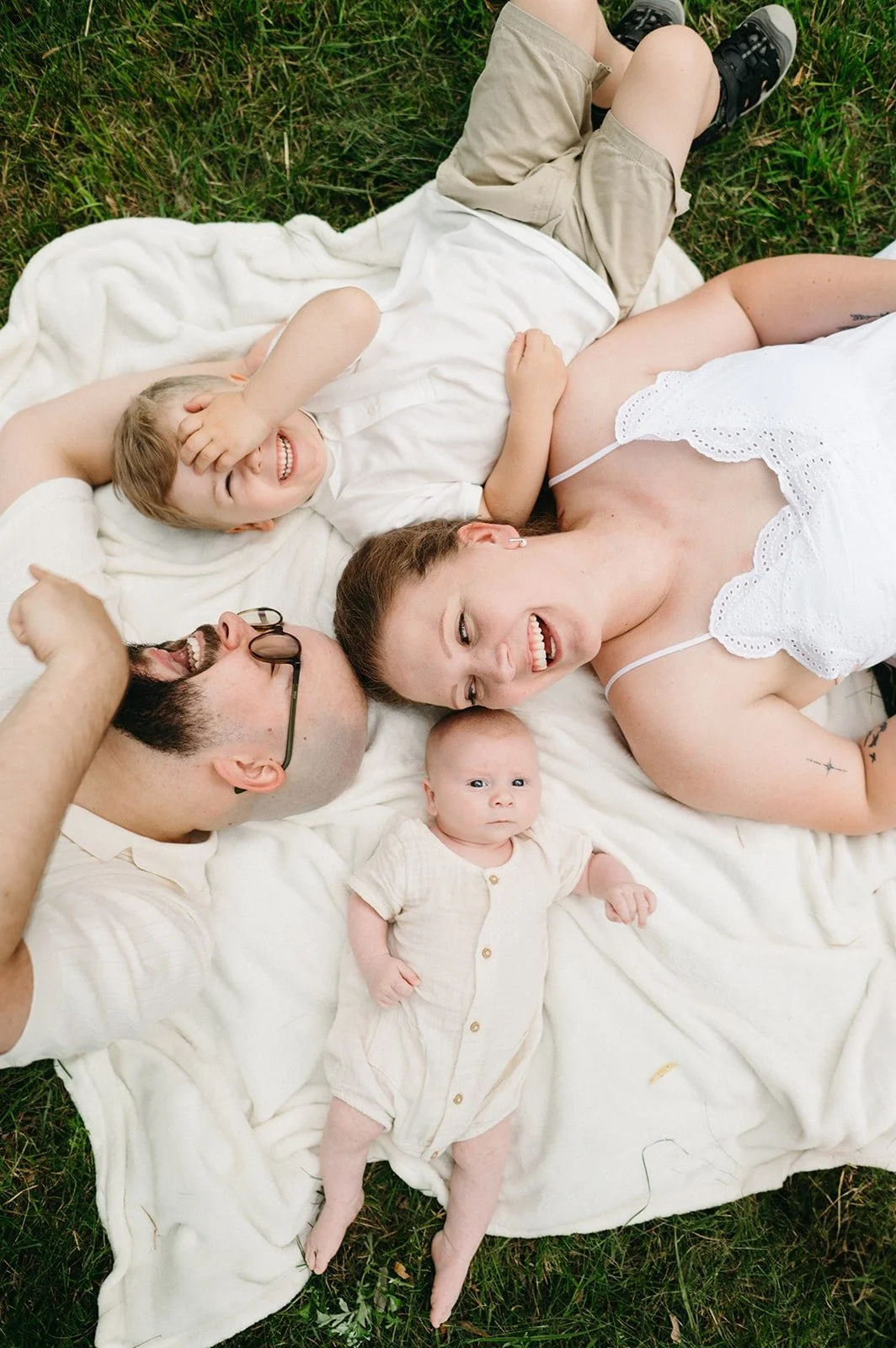 Famille Vincent Labonté assise sur une couverture blanche dans l’herbe verte, composée d’une femme, d’un homme, de deux enfants et d’un bébé, tous souriants et visiblement amusés, captés par un Photographe mariage Québec.