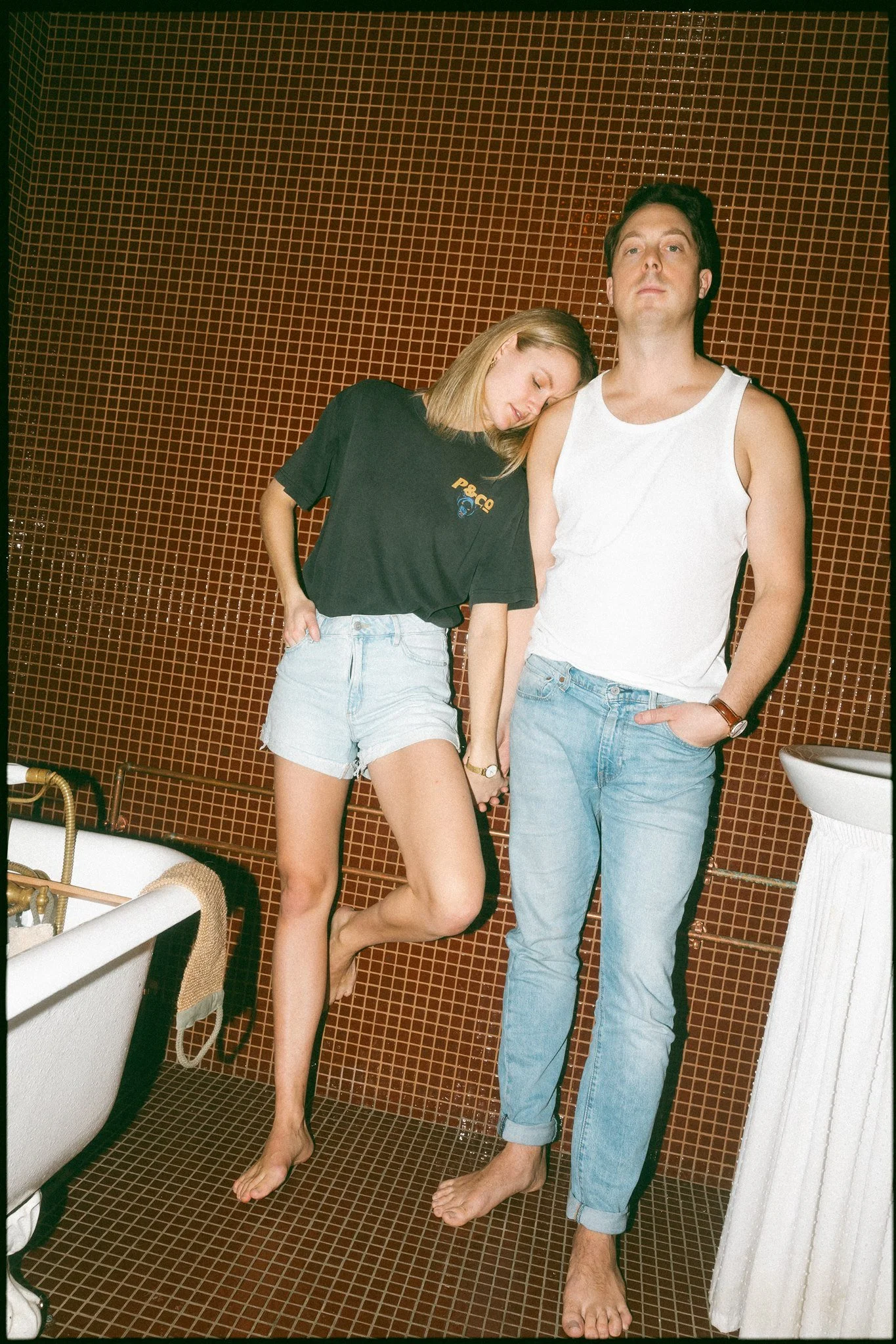 Deux personnes, un homme et une femme, debout dans une salle de bain avec un mur en carreaux marron, l'homme porte un débardeur blanc et un jean, la femme porte un t-shirt noir et un short en jean. La femme a la tête appuyée sur l'épaule de l'homme, 