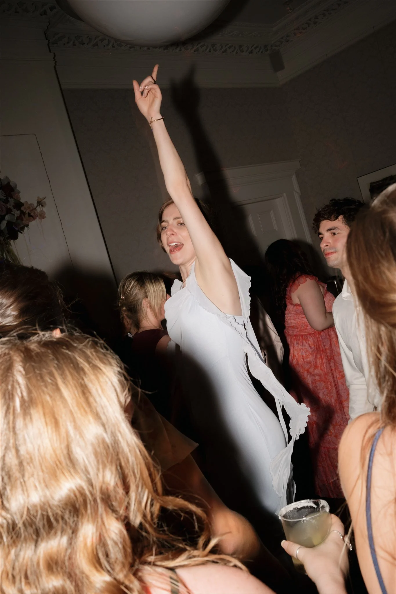 Une femme danse avec un bras levé lors d'une fête ou d'une célébration, entourée d'autres personnes dans une pièce élégante.