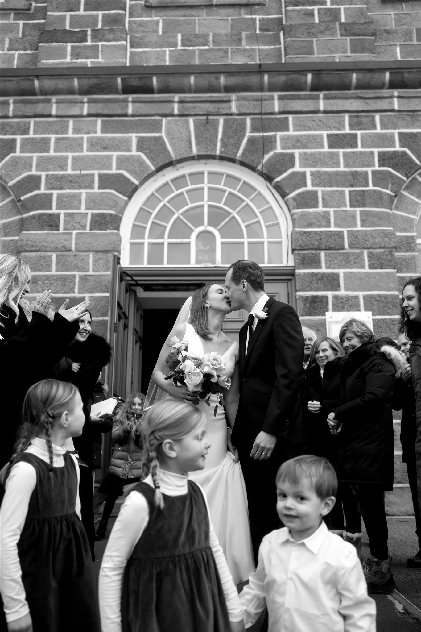 Un couple de mariés s'embrassant devant une groupe de personnes, avec des enfants en premier plan, devant une façade en briques avec une fenêtre arquée. La scène est en noir et blanc.