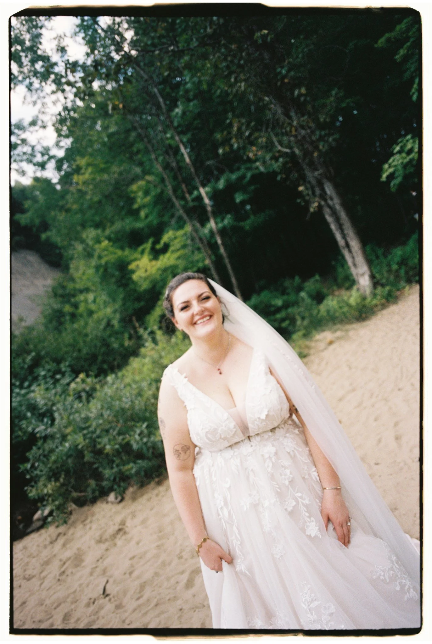 Une femme souriante portant une robe de mariage blanche avec des détails en dentelle, dans un environnement extérieur avec des arbres verts en arrière-plan.