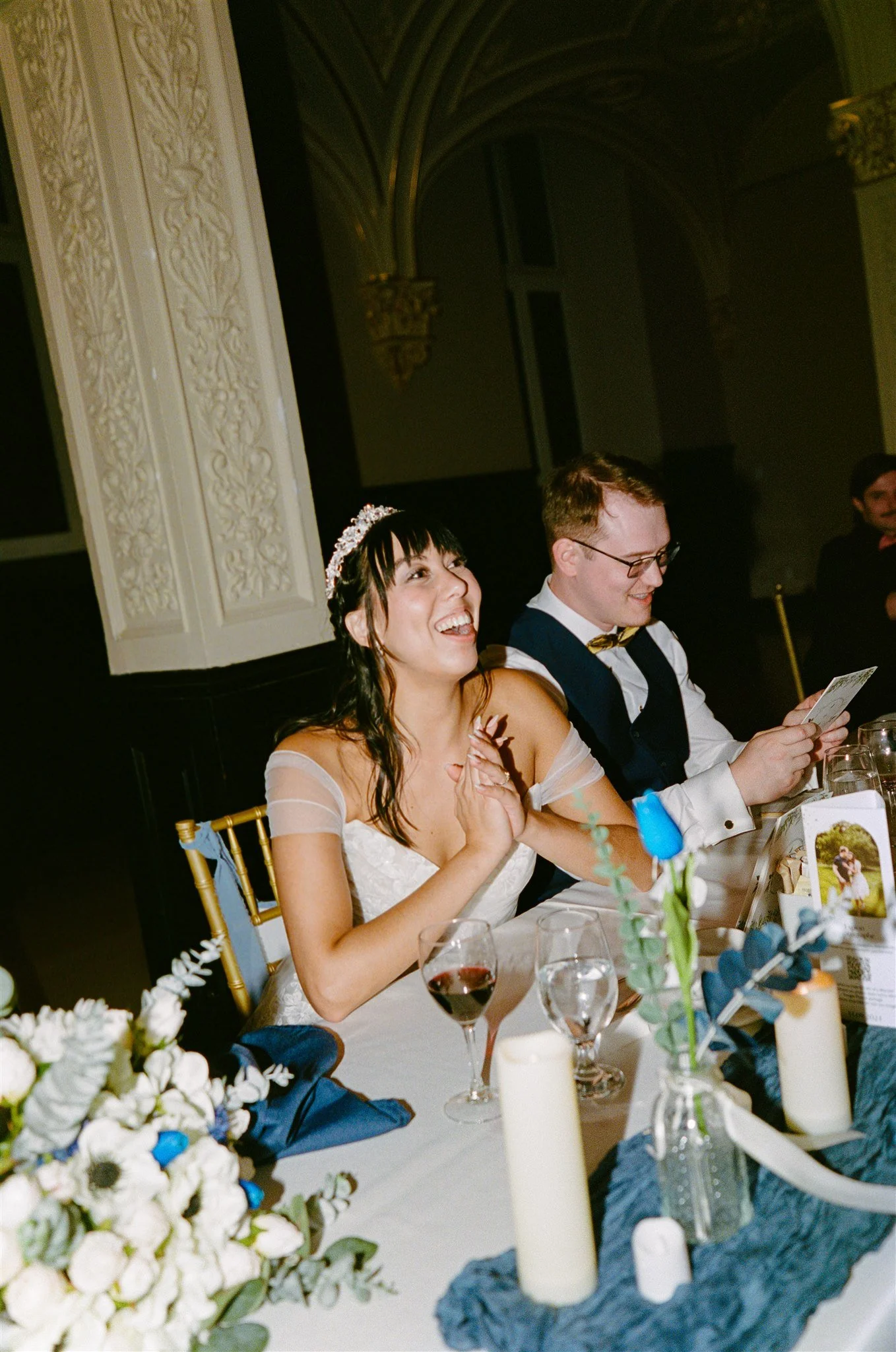Un couple de mariés souriant assis à une table lors d'une réception de mariage, entouré de décorations florales et de bougies.
