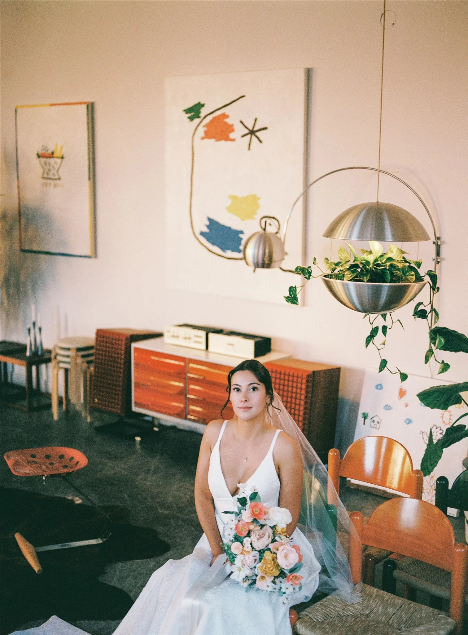 Une jeune femme en robe blanche avec un voile de mariée, tenant un bouquet de fleurs colorées, assise dans une pièce décorée de tableaux et de meubles en bois.