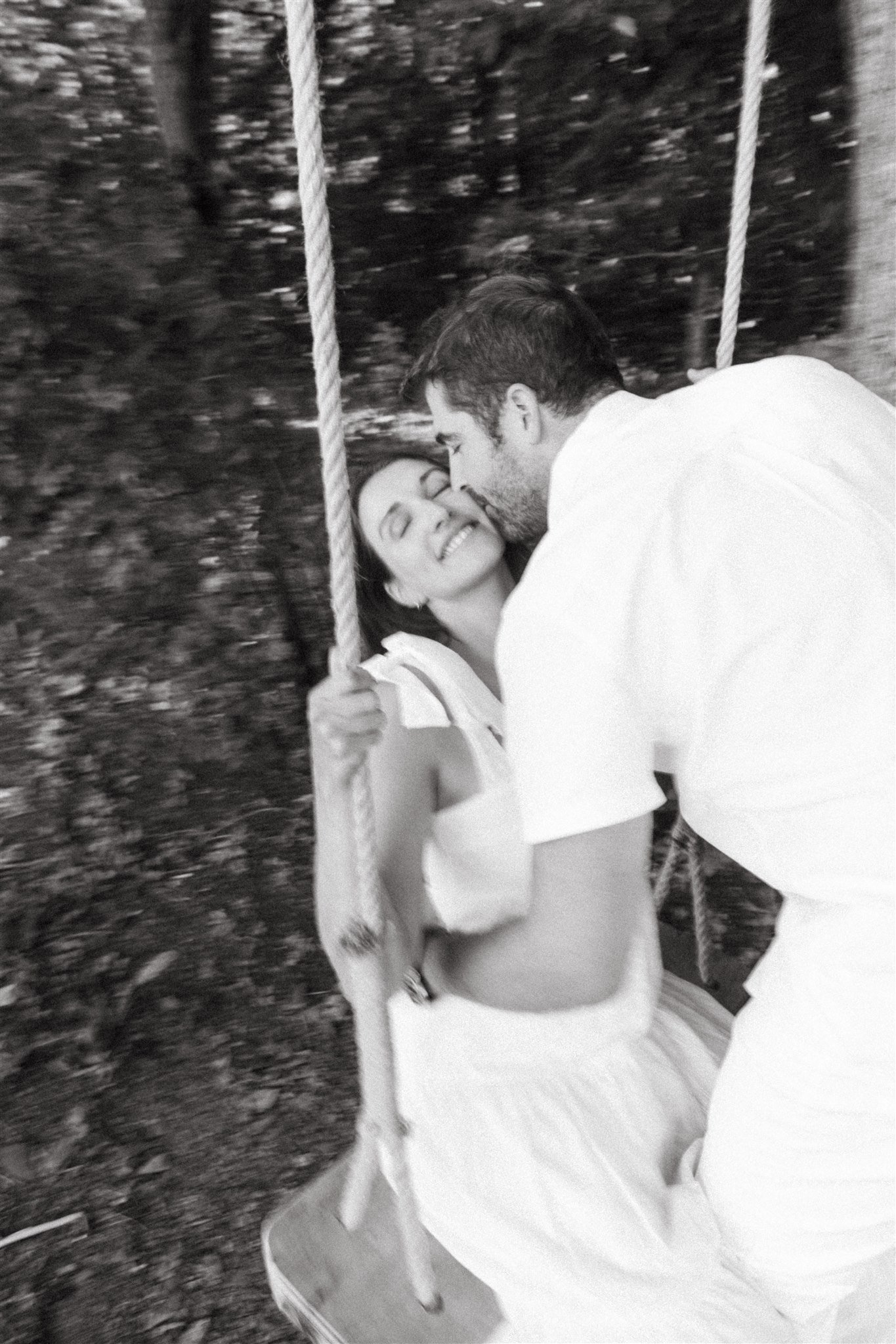 Un couple échange un baiser devant une balançoire en bois dans un environnement naturel, en noir et blanc.