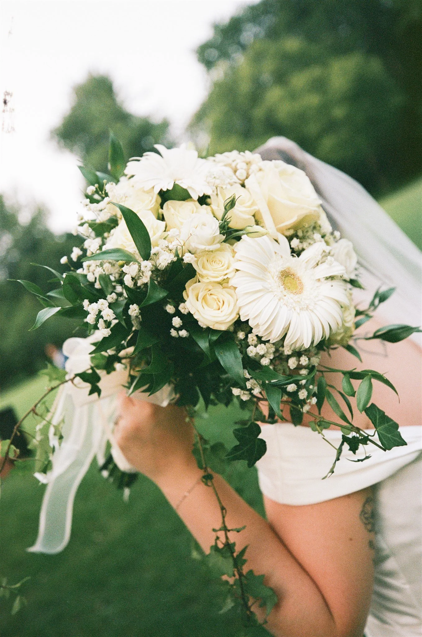 Poignée de main d'une personne tenant un bouquet de fleurs blanches, notamment des roses et des gerberas, avec un fond flou de nature.