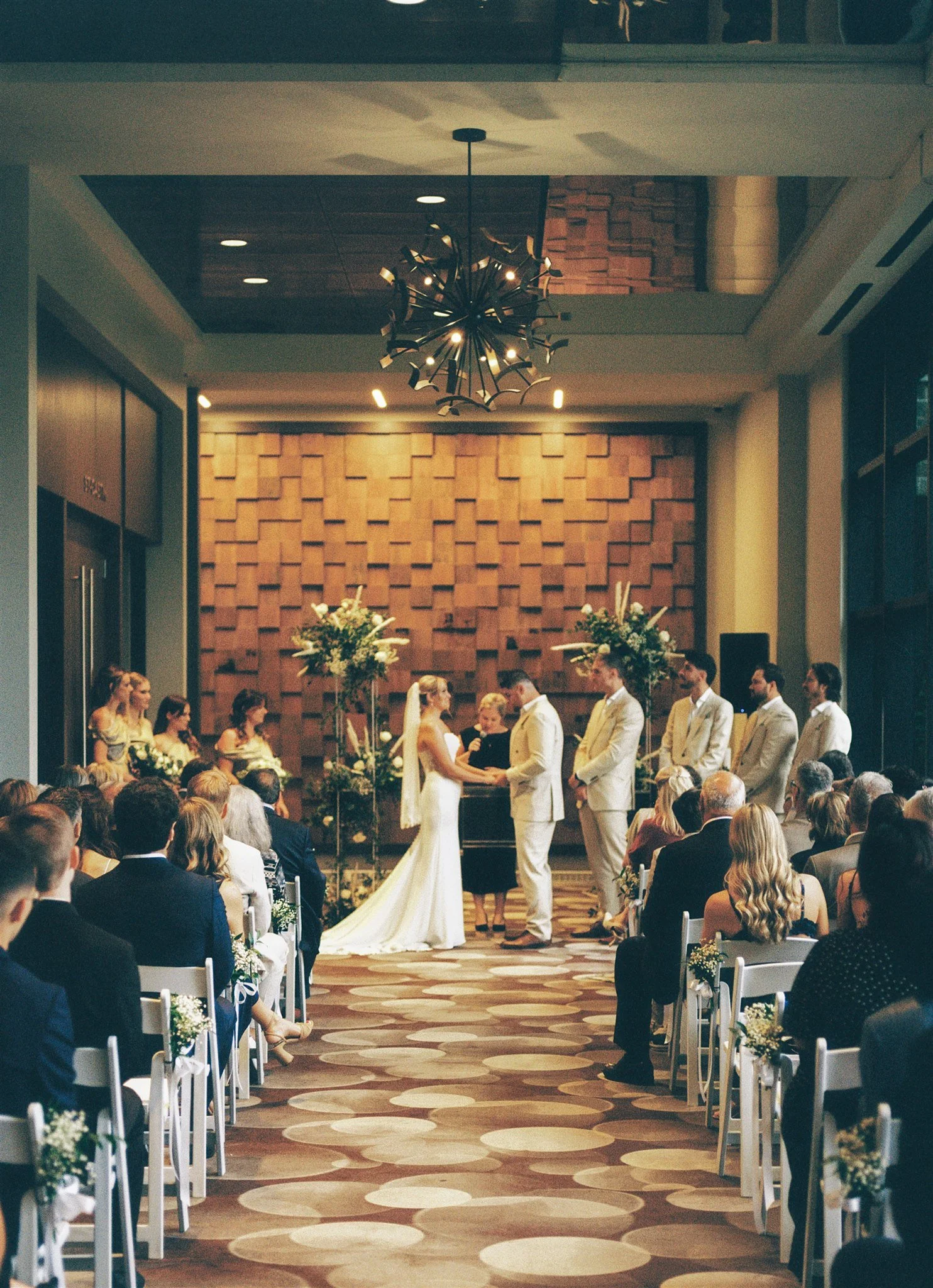 Une cérémonie de mariage se déroule dans une salle élégante avec un mur en bois et des décorations florales. Le couple de mariés se tient devant l'officiant, entouré de témoins. Les invités sont assis et regardent la cérémonie.