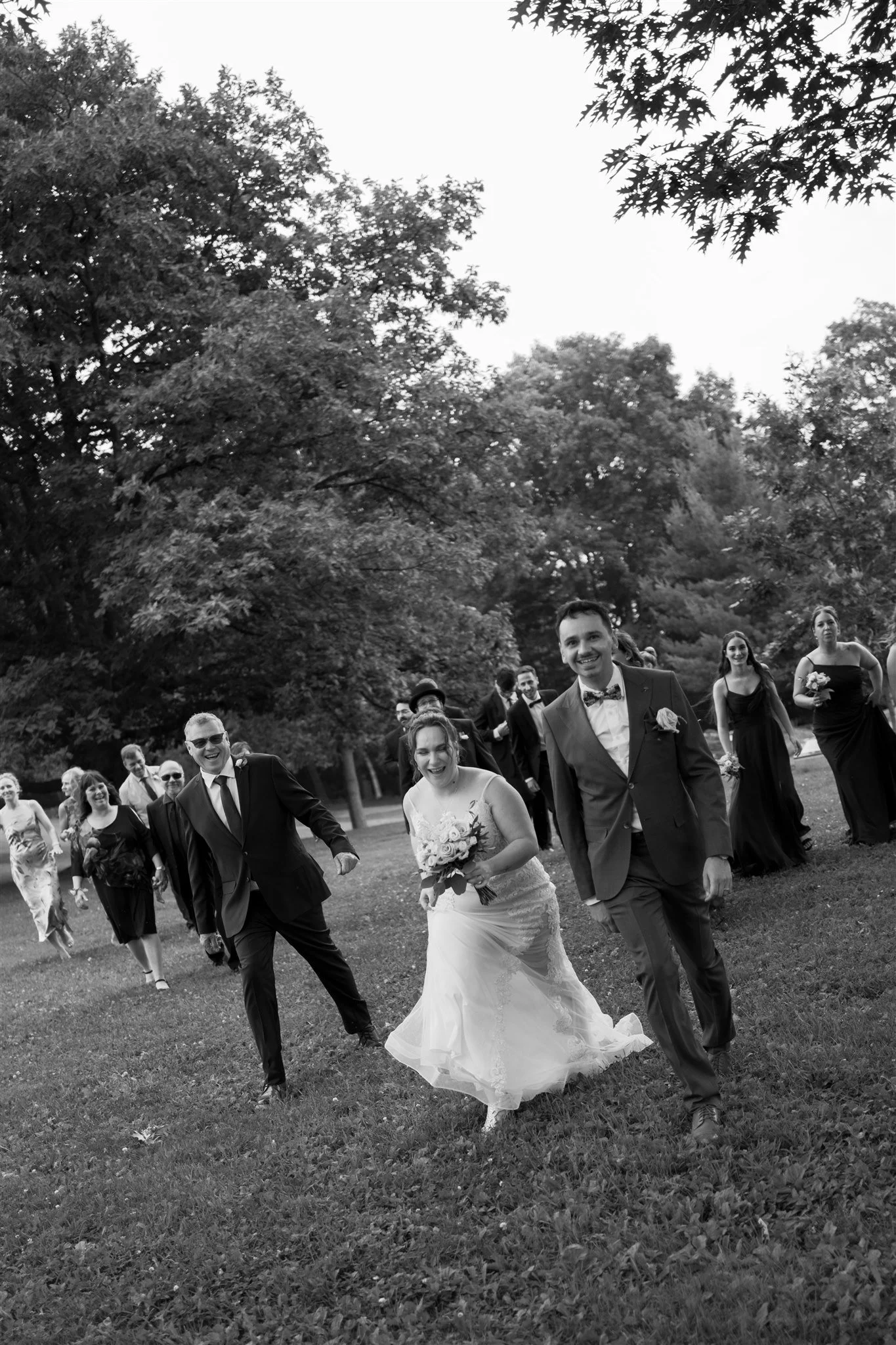 Un groupe de personnes en costume de mariage et de soirée se promènent dans un parc vert avec des arbres en arrière-plan, lors d'un mariage.