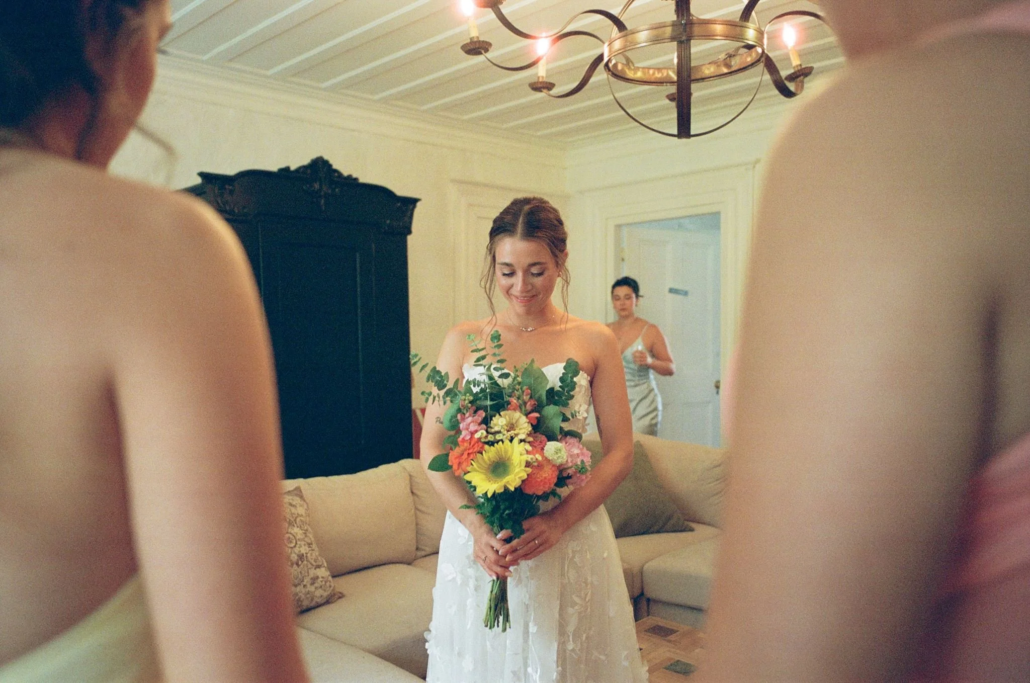 Femme en robe de mariée tenant un bouquet de fleurs dans un salon, entourée de personnes floues lors de la préparation pour un mariage.