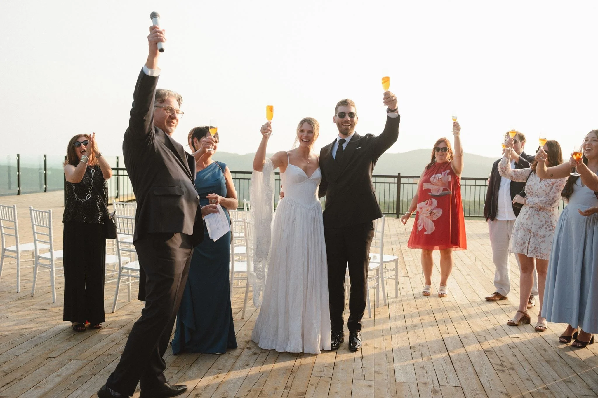 Groupe de personnes lors d'une célébration de mariage en plein air, avec un couple marié au centre. Les invités portent des tenues élégantes et tiennent des verres orange-rouge à garniture d'ananas, tous souriants et joyeux.