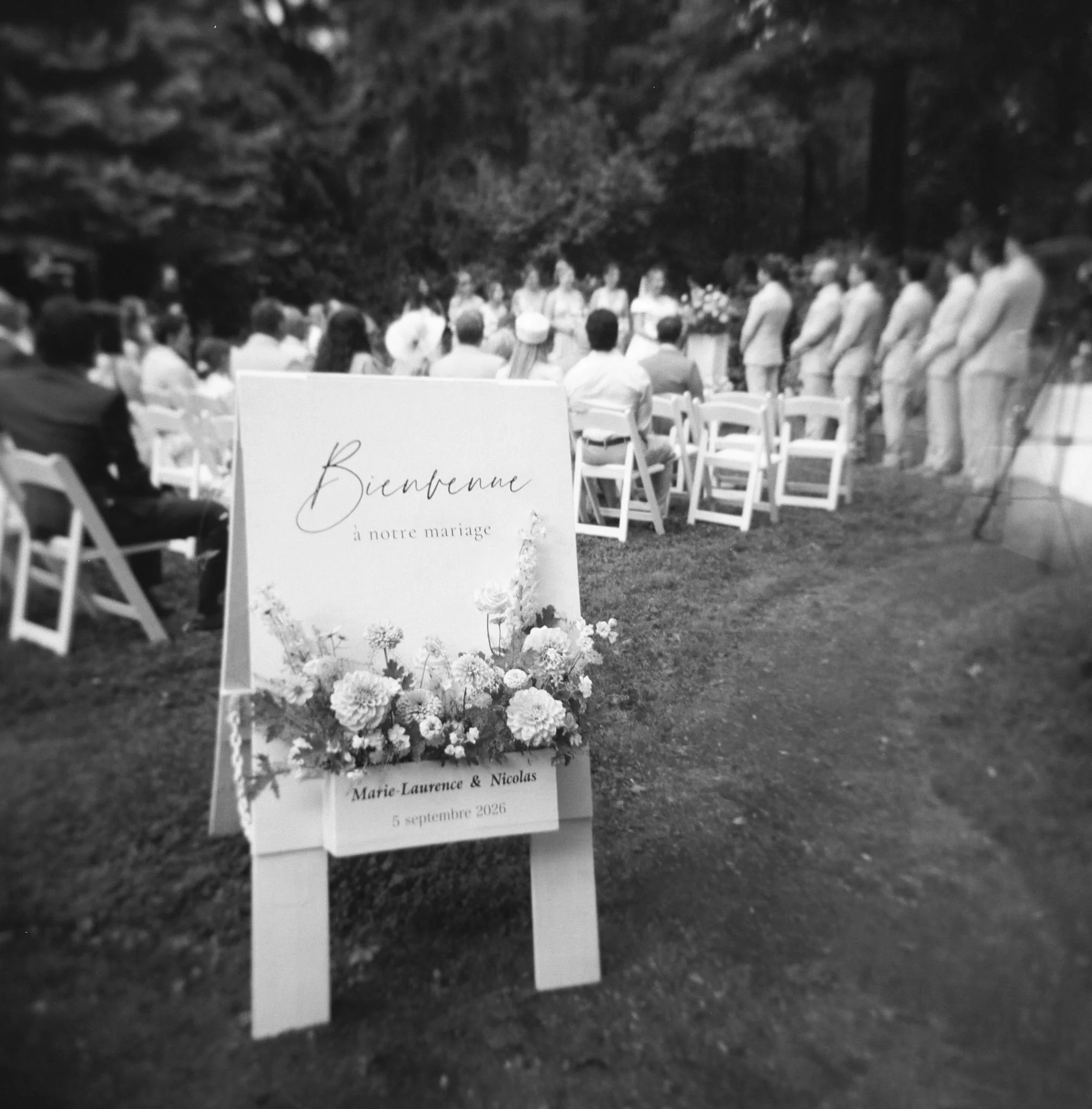 Un panneau en bois avec la mention 'Bienvenue à notre mariage' entouré de fleurs, dans un cadre extérieur lors d'une cérémonie célébrée en plein air.