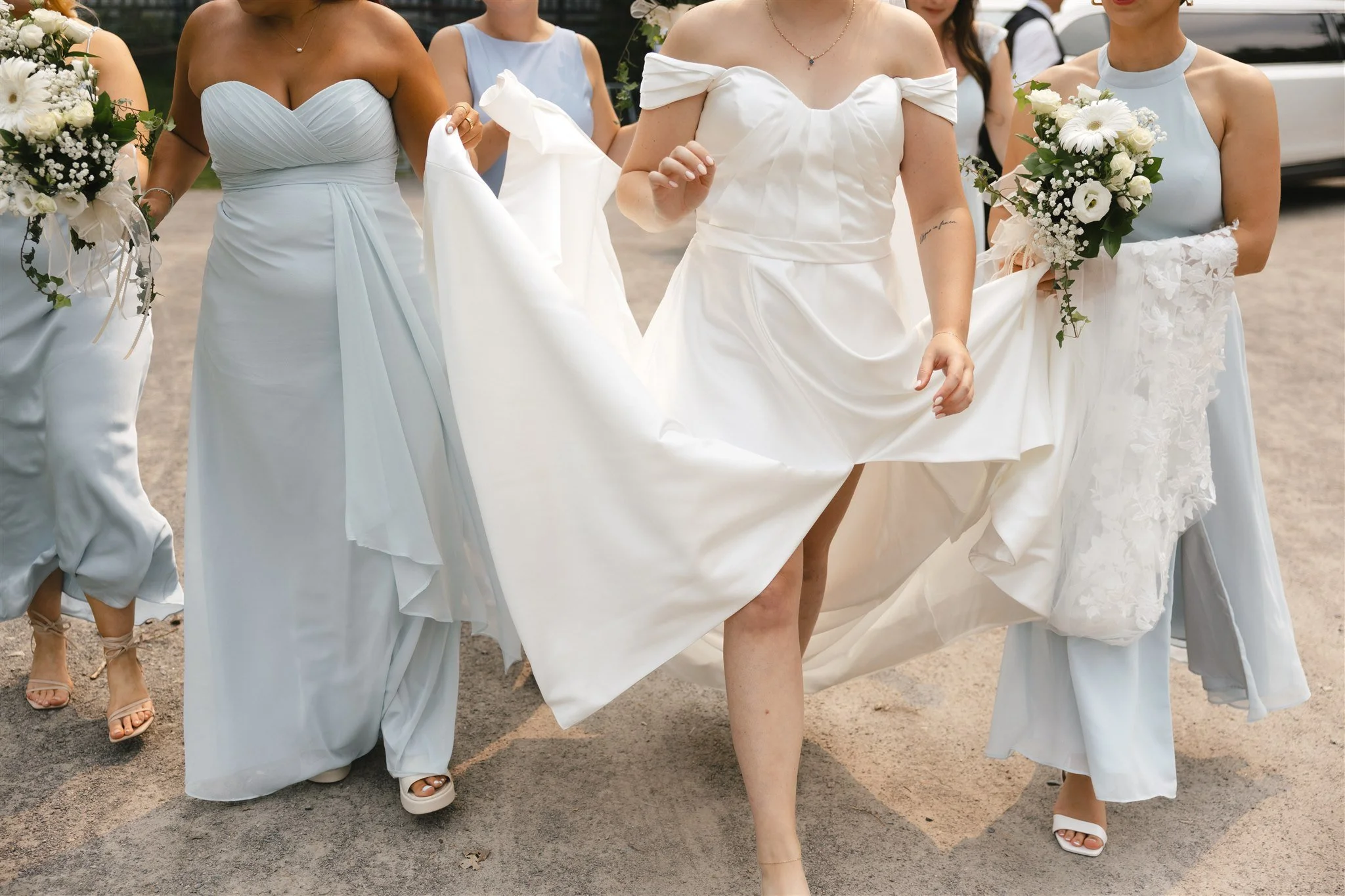 Femmes en robes de mariée ou de soirée blanche, participant à un mariage ou un événement festif, portant des bouquets de fleurs, marchant sur une surface en terre.