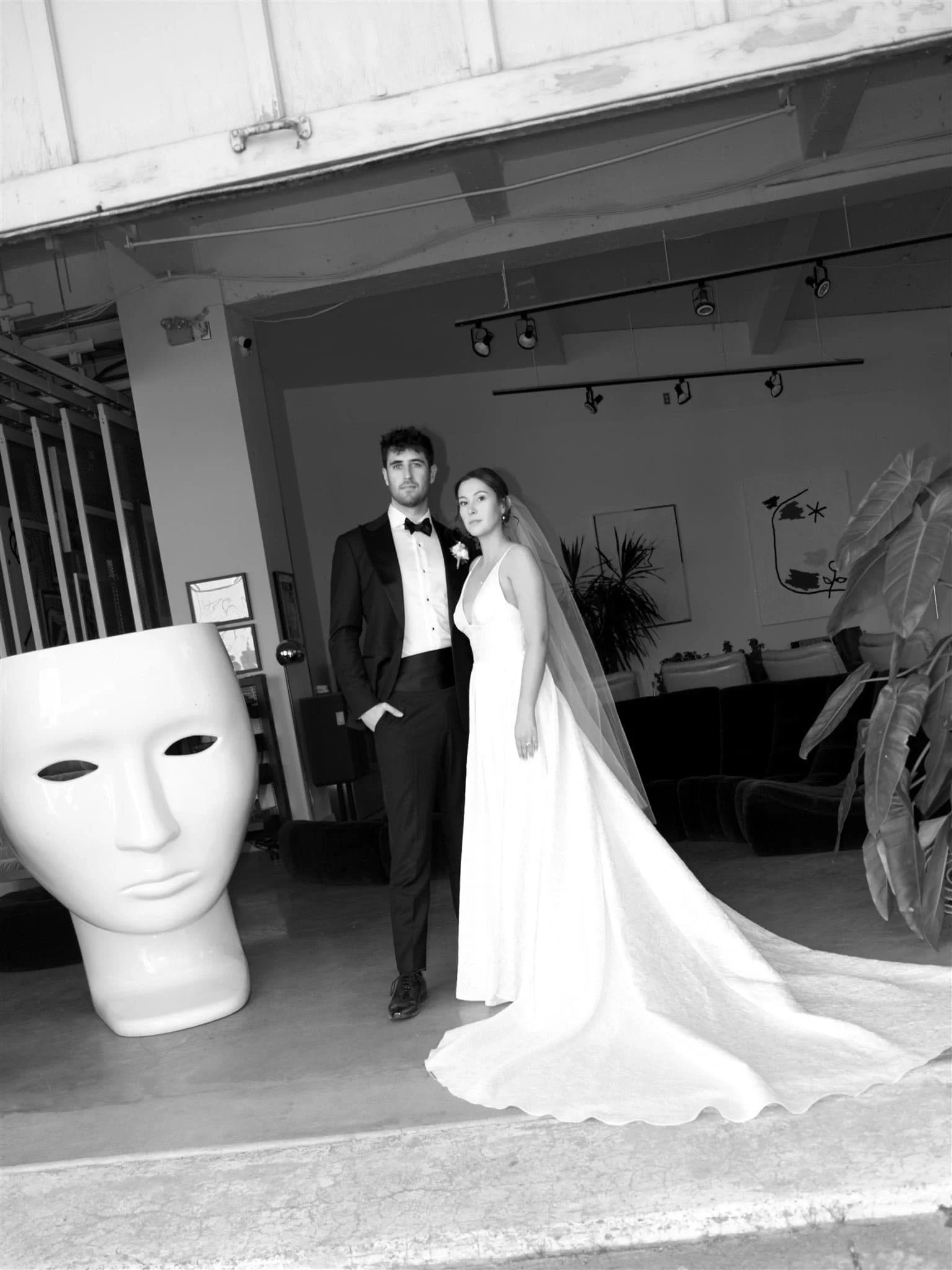 Homme en costume noir avec nœud papillon et femme en robe de mariage longue avec voile posant dans un décor intérieur contemporain orné de plantes et d’un grand masque blanc, captés par un Photographe mariage Québec  dans un commerce du Vieux Québec.