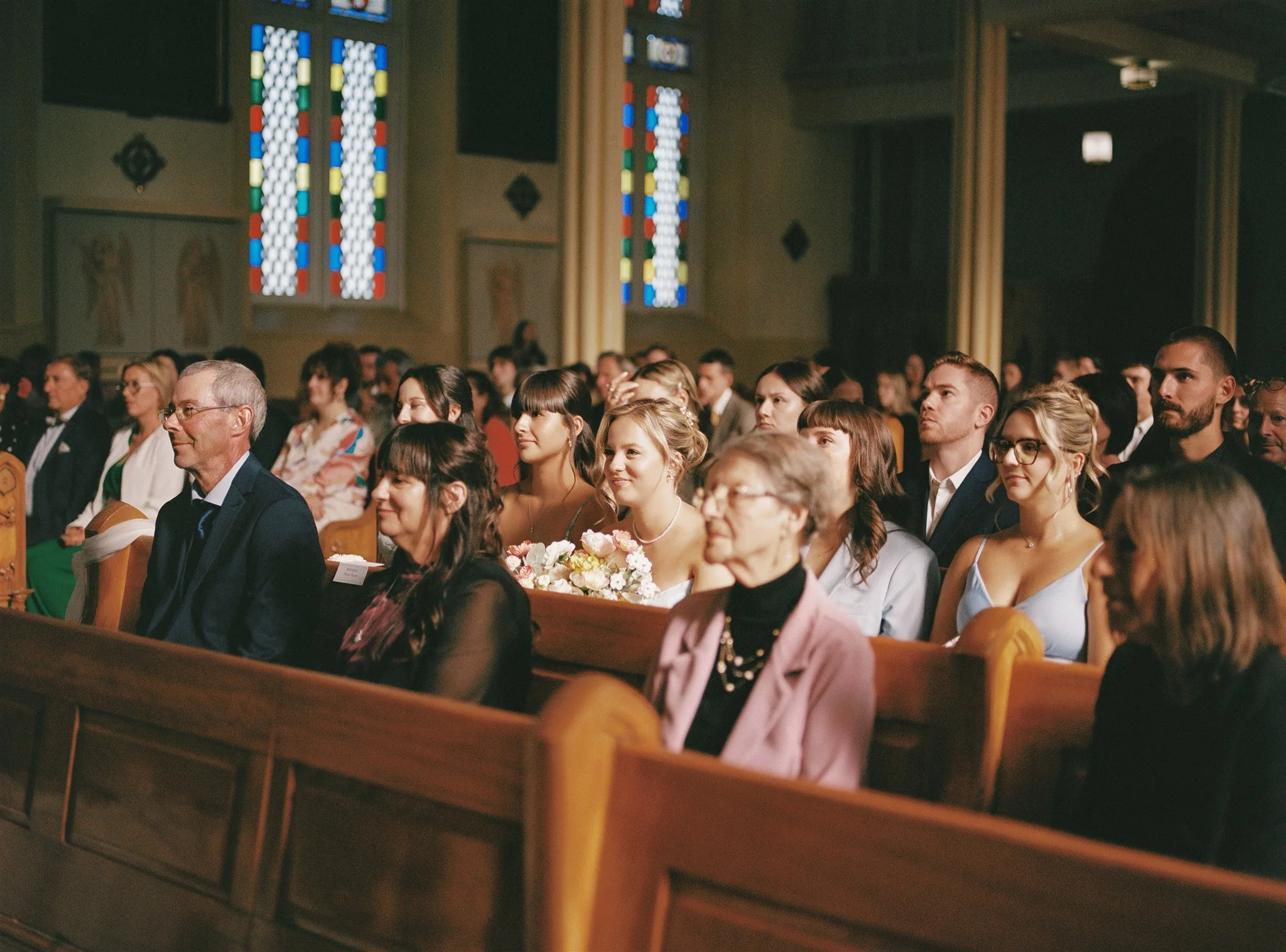 Une assemblée d'hommes et de femmes assis dans une église, lors d'une cérémonie ou d'un événement religieux, avec des fenêtres colorées en arrière-plan.