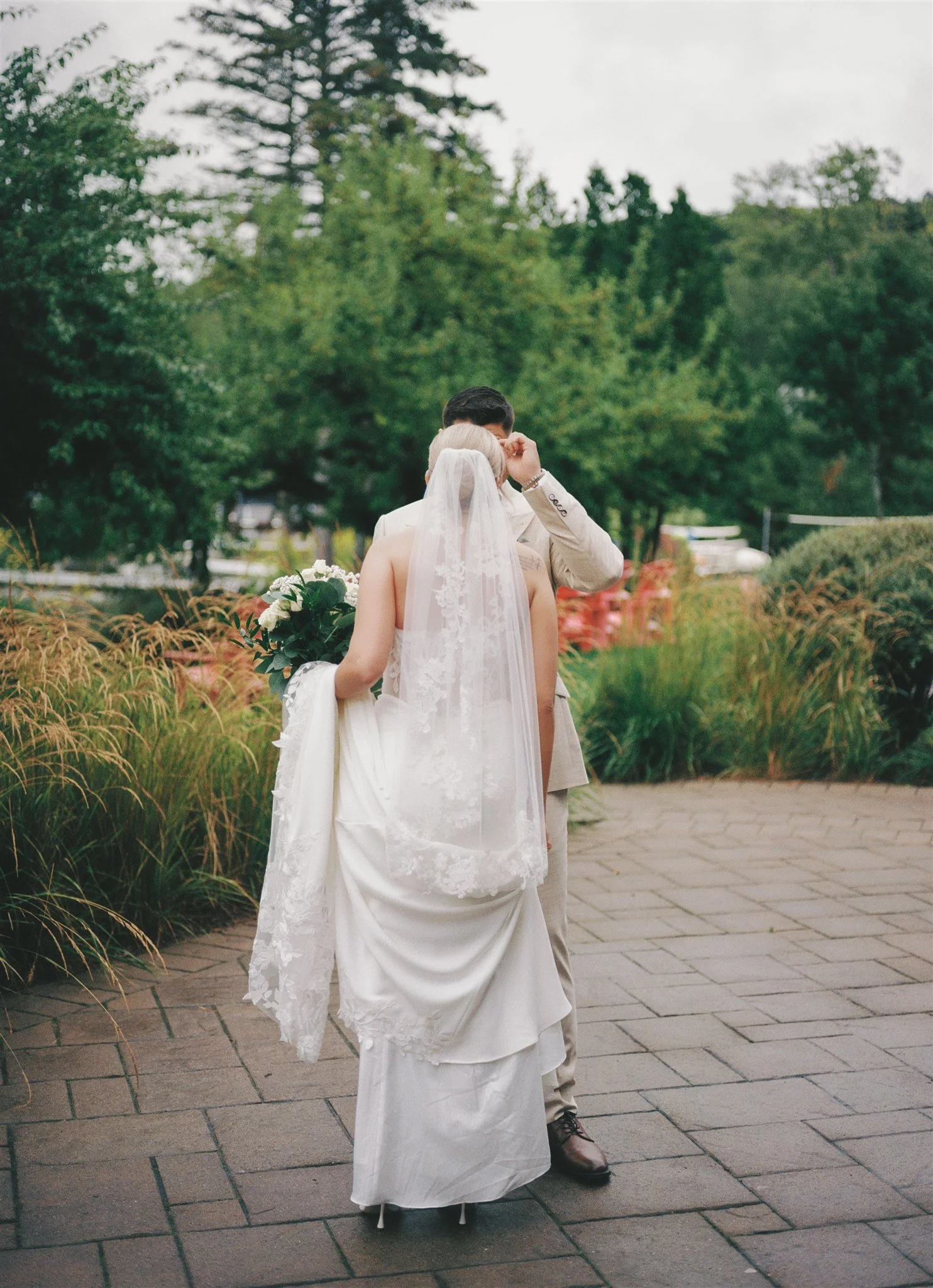 Un couple de mariés lors d'une cérémonie en plein air sous un ciel nuageux, la mariée porte une robe blanche avec une voilette et tient un bouquet de fleurs, l'homme porte un costume beige.