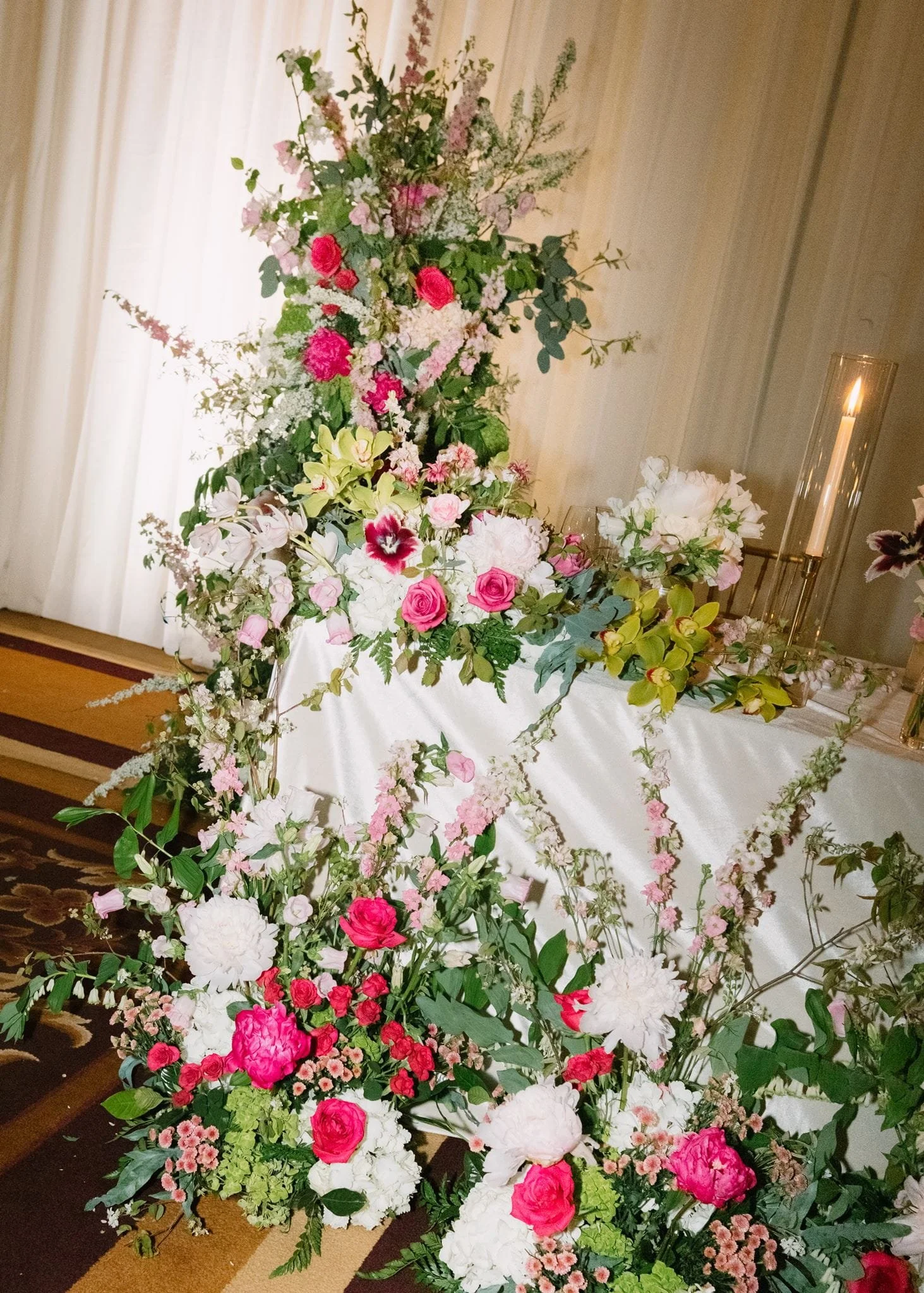Grand arrangement floral avec roses roses, hortensias blancs, et autres fleurs colorées, décorant une table lors d'un événement, avec une bougie allumée en arrière-plan.