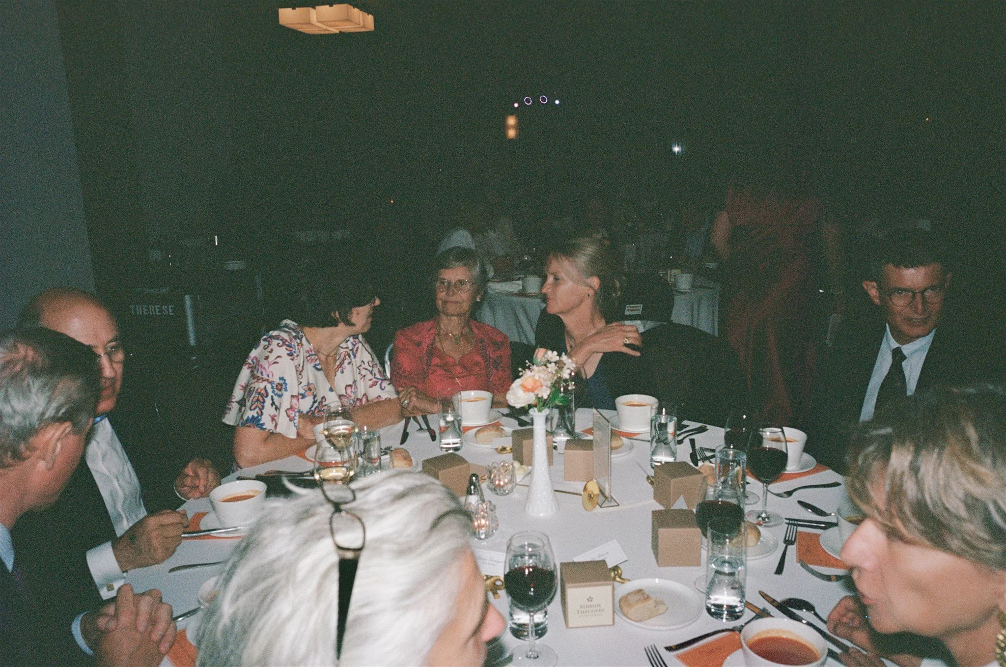 Groupe de personnes assises autour d'une table lors d'un dîner ou d'une célébration, avec des plats, des verres de vin et des fleurs, dans un cadre intérieur
