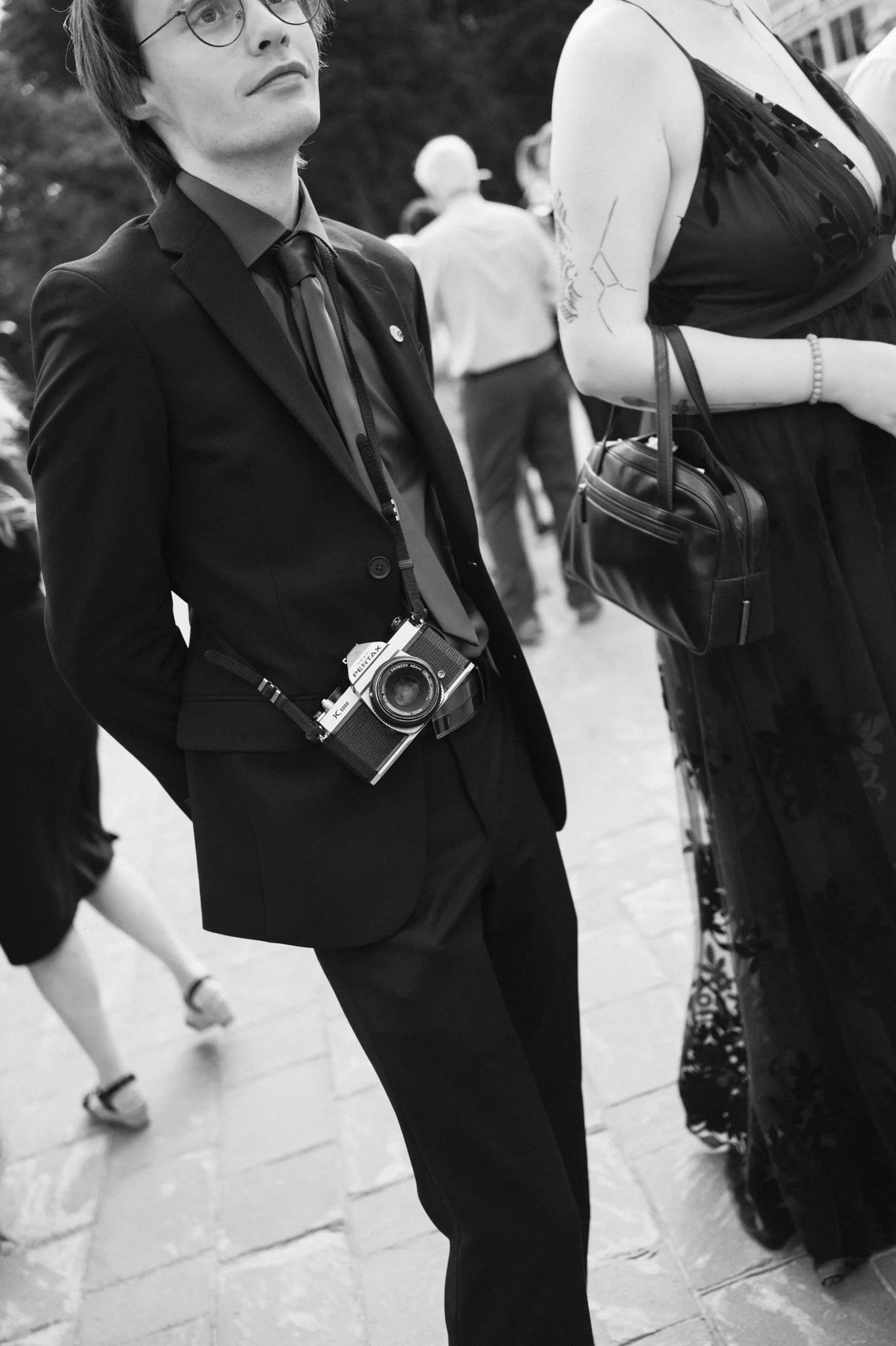 Une personne masculine portant un costume noir, des lunettes et un appareil photo accroché à la taille marche dans une foule lors d'un événement en plein air.