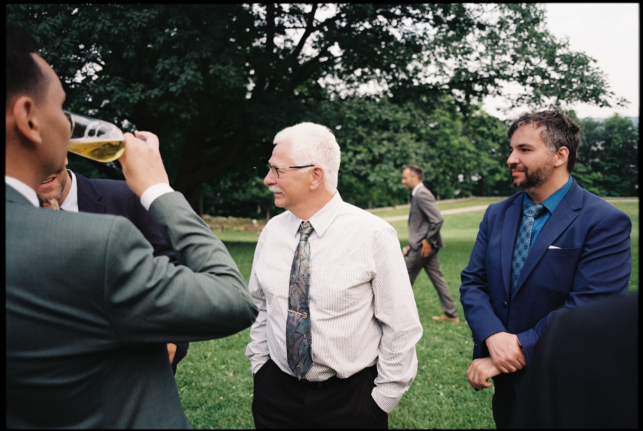 Groupe d'hommes en costume discutant et buvant lors d'une rencontre en plein air dans un parc avec beaucoup d'arbres.