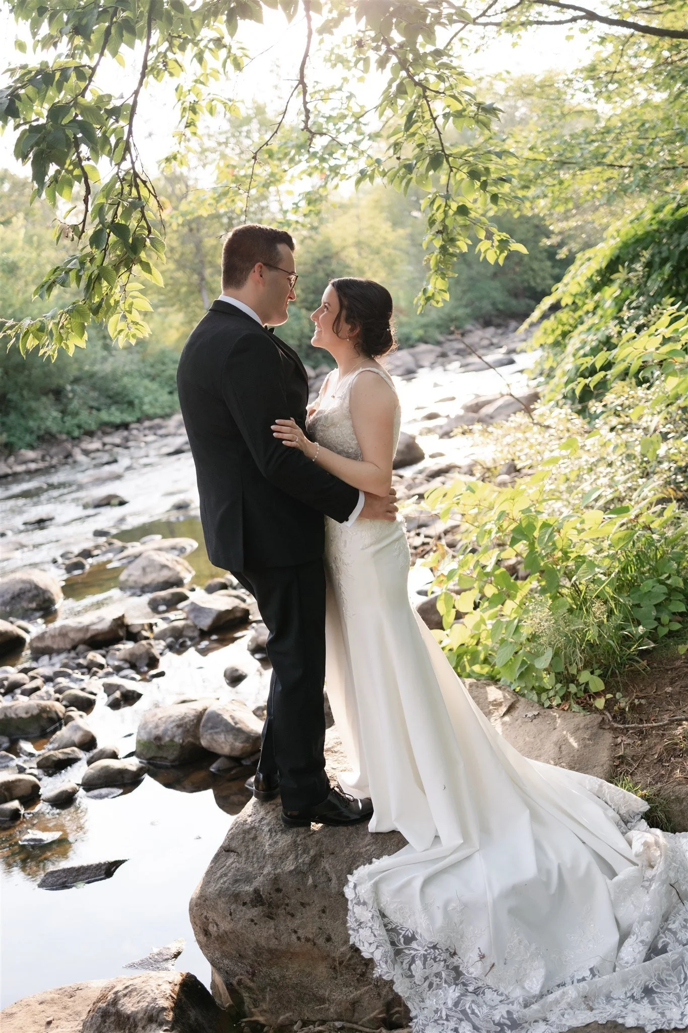 Un couple marié en robe de mariée et costume élégants se tiennent sur un rocher près d'une rivière dans la nature, sous un arbre avec de la verdure, lors d'une séance photo de mariage en plein air.