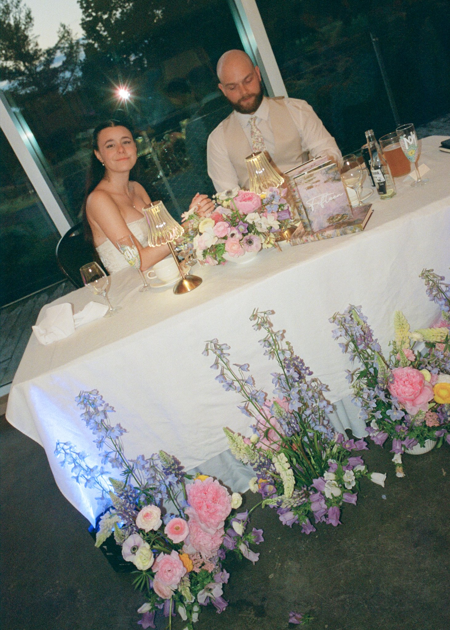Table de mariage décorée avec un centre de fleurs, des lampes de table, des verres et des menus, dans une salle avec fenêtres derrière, deux personnes assises à la table, une femme et un homme