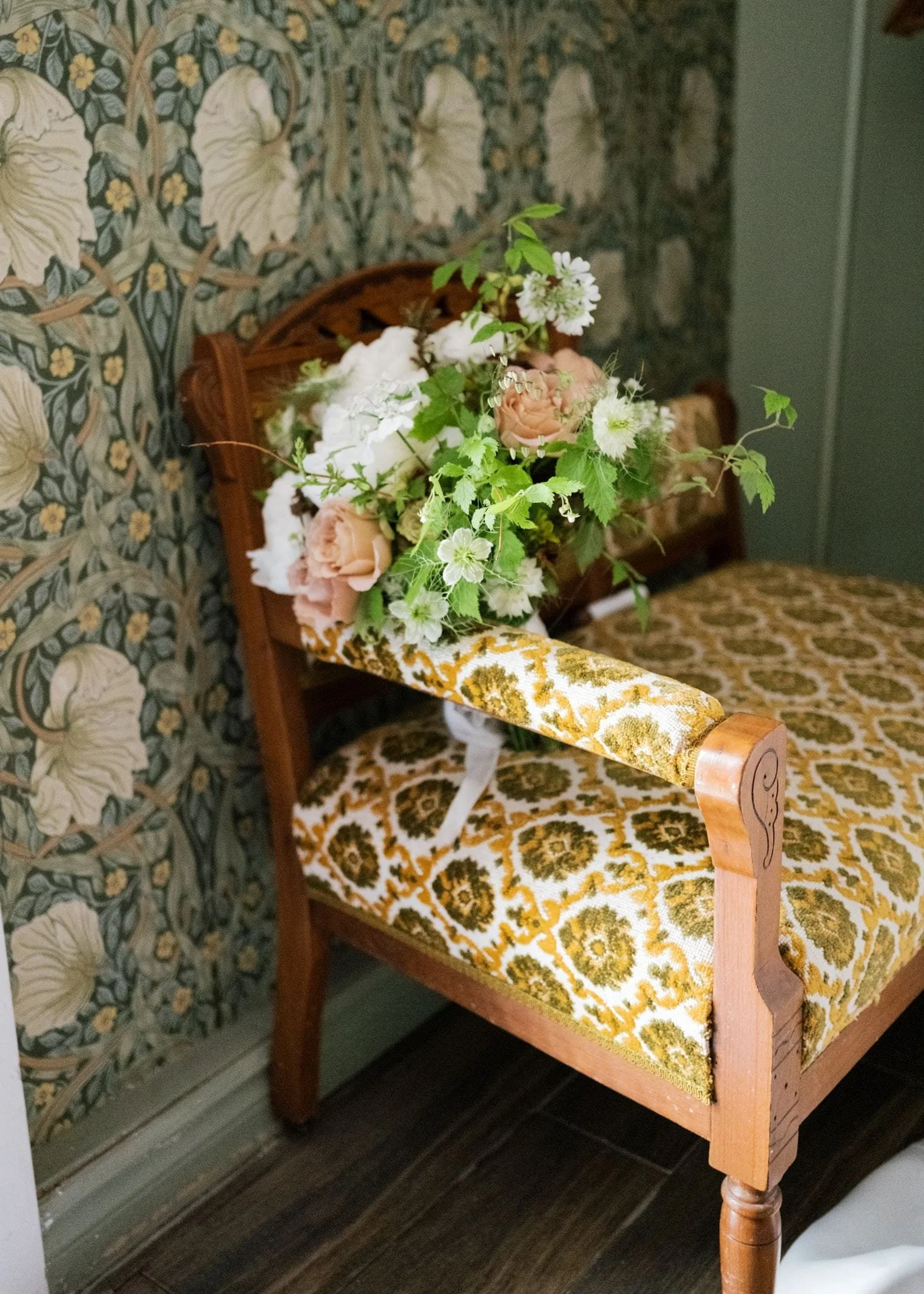 Une chaise ancienne en bois avec un coussin en tissu à motif jaune et blanc, un bouquet de fleurs colorées est posé dessus, mur à motifs floraux en arrière-plan.
