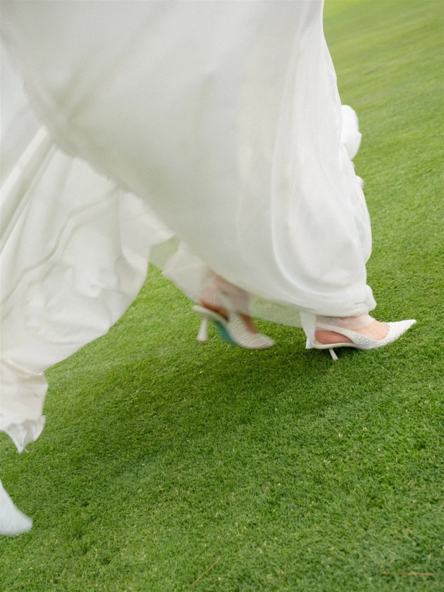 Une personne portant des chaussures à talons blancs avec des semelles brillantes, marchant sur une surface de pelouse verte, avec une robe blanche fluide et un chapeau blanc en tissu léger.
