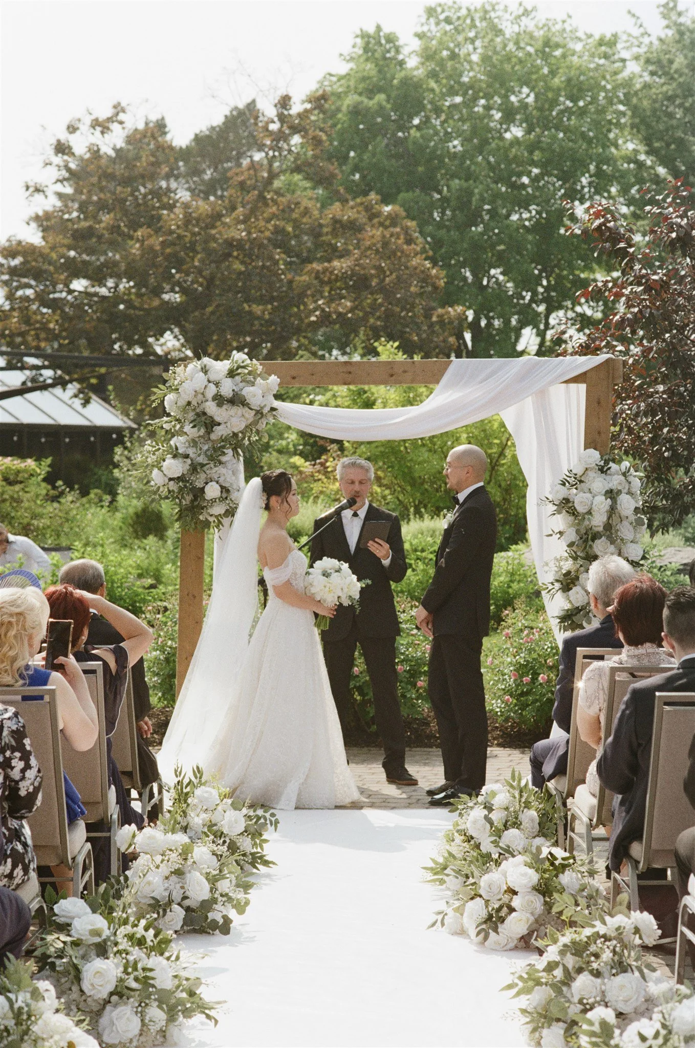 Un couple en mariage échange leurs vœux lors d'une cérémonie en extérieur, sous une arche décorée de fleurs blanches, entourée de famille et d'amis.
