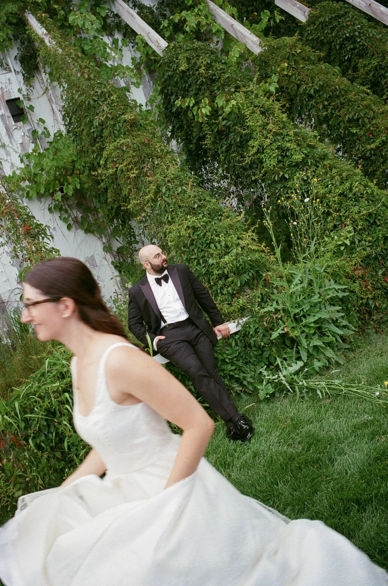 Une femme en robe blanche passe devant une personne en smoking qui est assise sur un banc dans un jardin verdoyant.