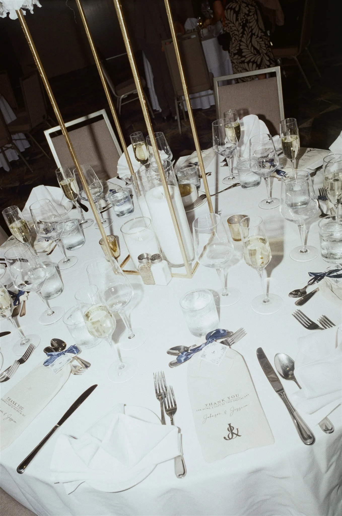 Table de banquet ornée de verres à vin, assiettes, couverts, verres d'eau, et un menu de remerciement. En arrière-plan, des invités et un décor de fête.