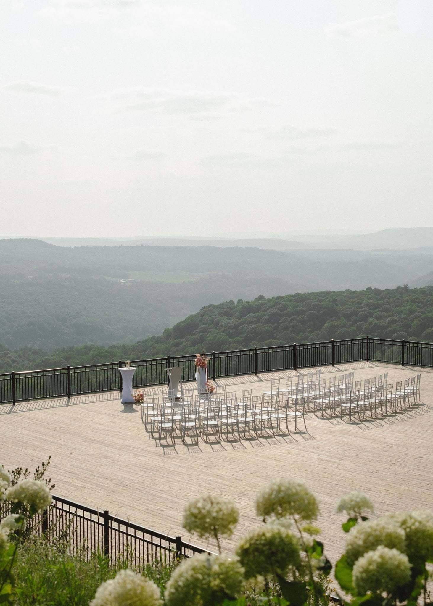 Une terrasse en plein air prête pour un événement, avec des chaises alignées, des tables avec décorations florales, surplombant un paysage de montagnes verdoyantes et un ciel légèrement nuageux.
