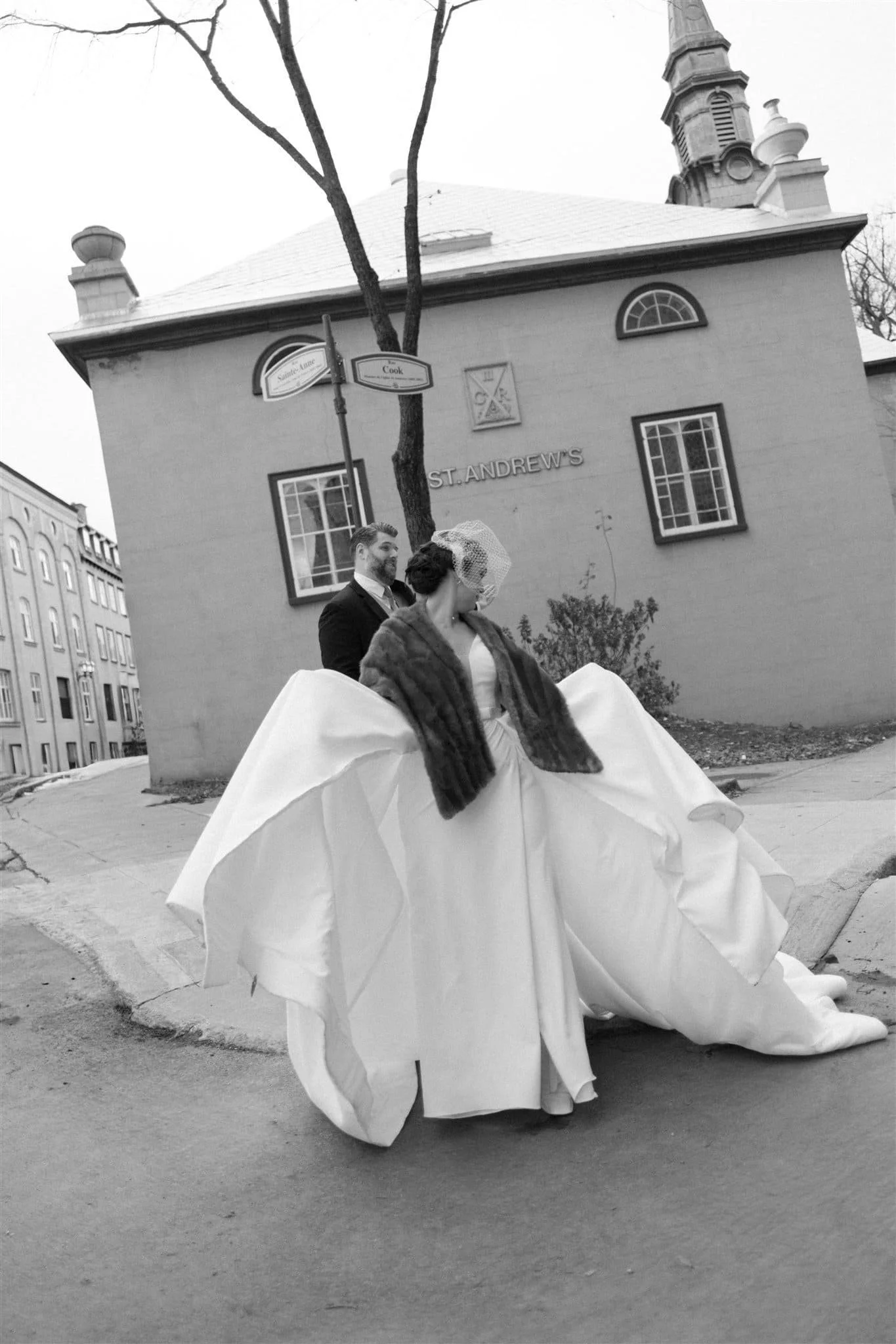 Femme en robe de mariée et homme en costume posant devant un bâtiment portant le nom St. Andrews, dans une rue du Vieux Québec en noir et blanc, captés par un Photographe mariage Québec.