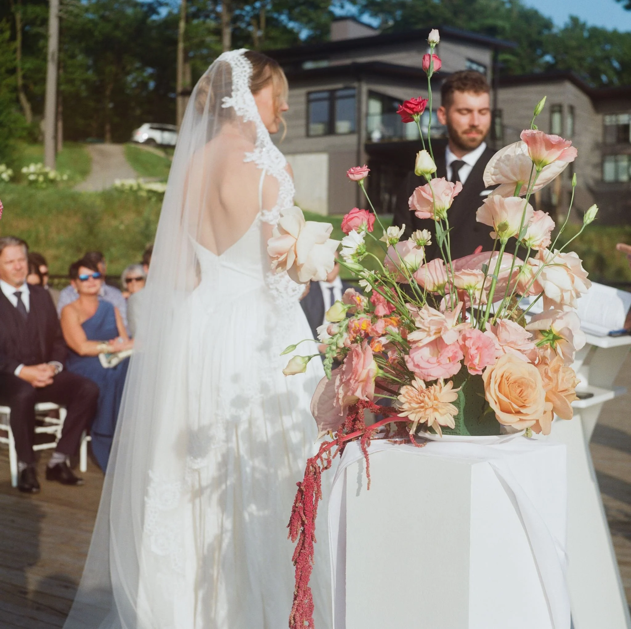 Un couple lors d'un mariage outdoors, avec la mariée en robe blanche avec voile et le marié en costume noir, devant une procession de personnes, en arrière-plan une maison moderne en bois, avec un panneau floral de fleurs roses, pêche, blanc et rouge