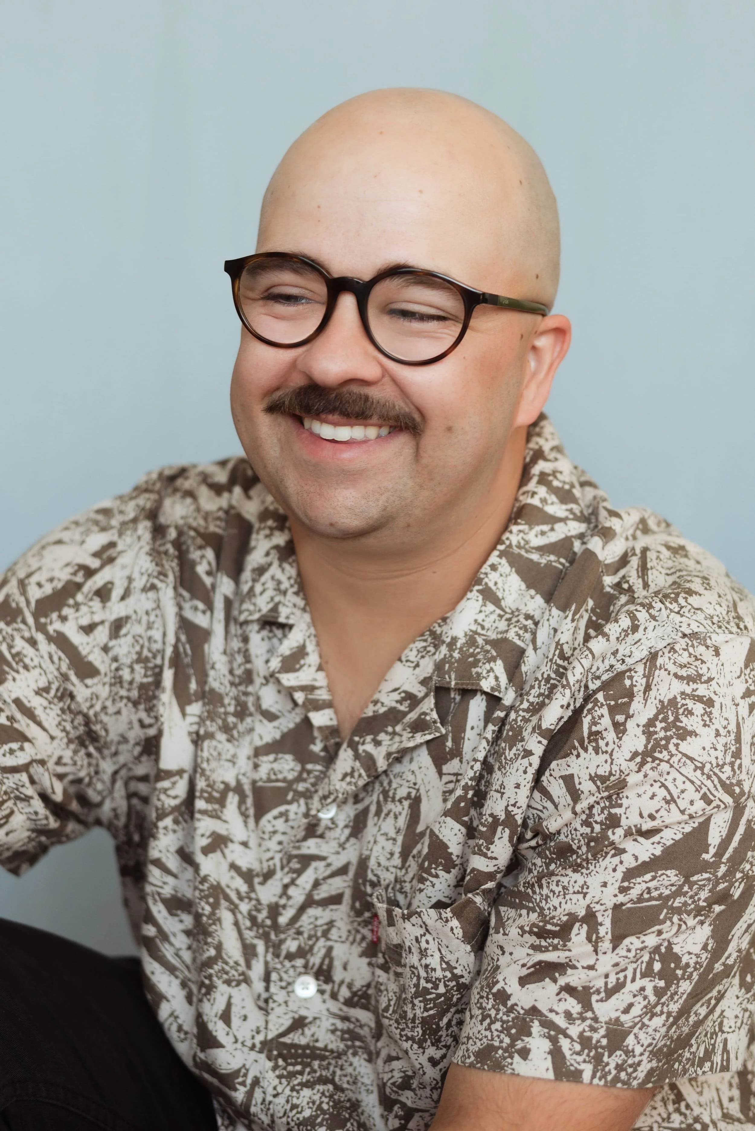 Un homme souriant portant des lunettes noires et une chemise à motifs marron et beige.