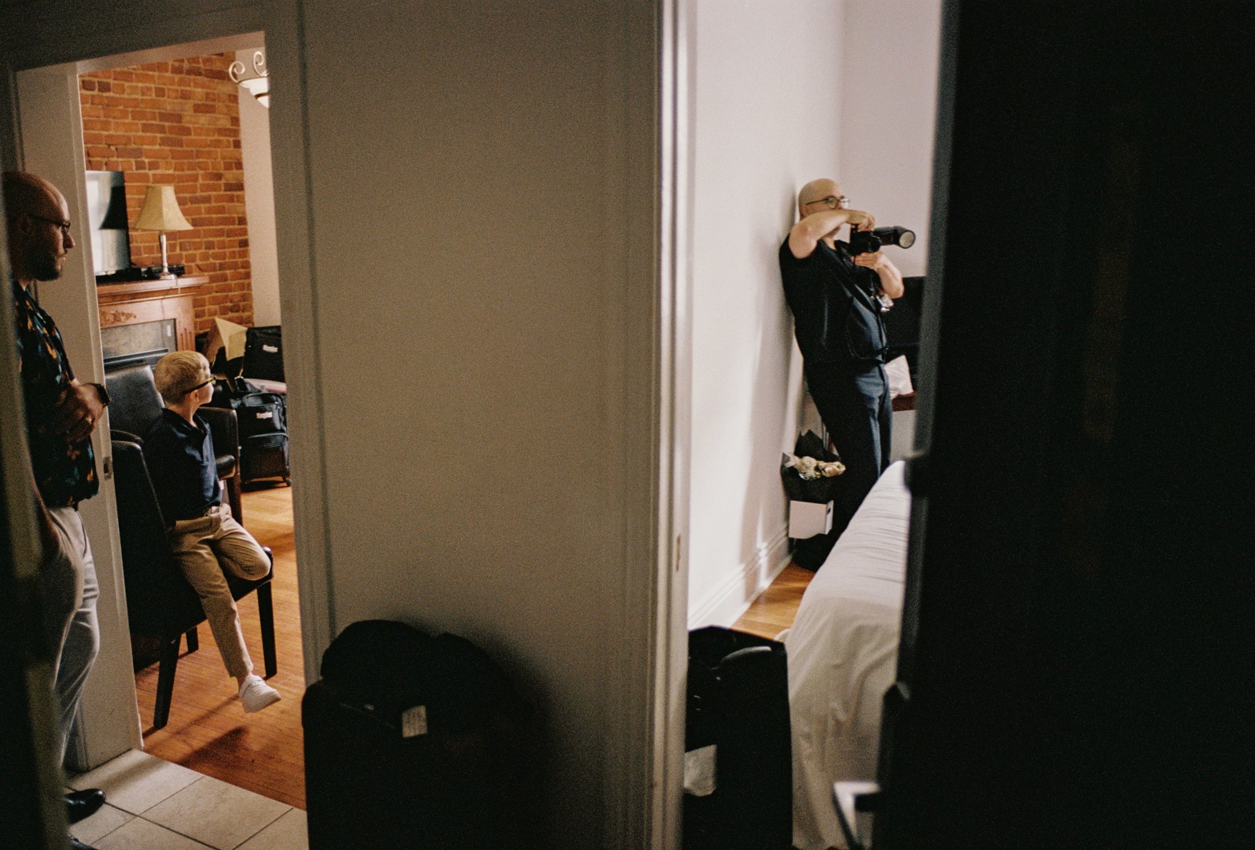 Scène dans un appartement avec deux personnes assises, l’une près d’une cheminée devant un mur de briques et l’autre prenant un selfie avec un smartphone, tandis qu’une troisième personne se tient à la porte en observant, le tout capté à travers un m