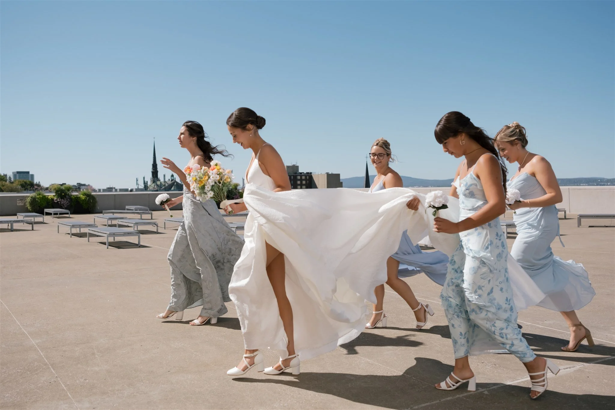 Un groupe de femmes habillées en robes de mariage blanches et bleues, avec des chaussures à talons, défilent sur une terrasse en plein air lors d'une journée ensoleillée, avec des bâtiments et un ciel bleu en arrière-plan.