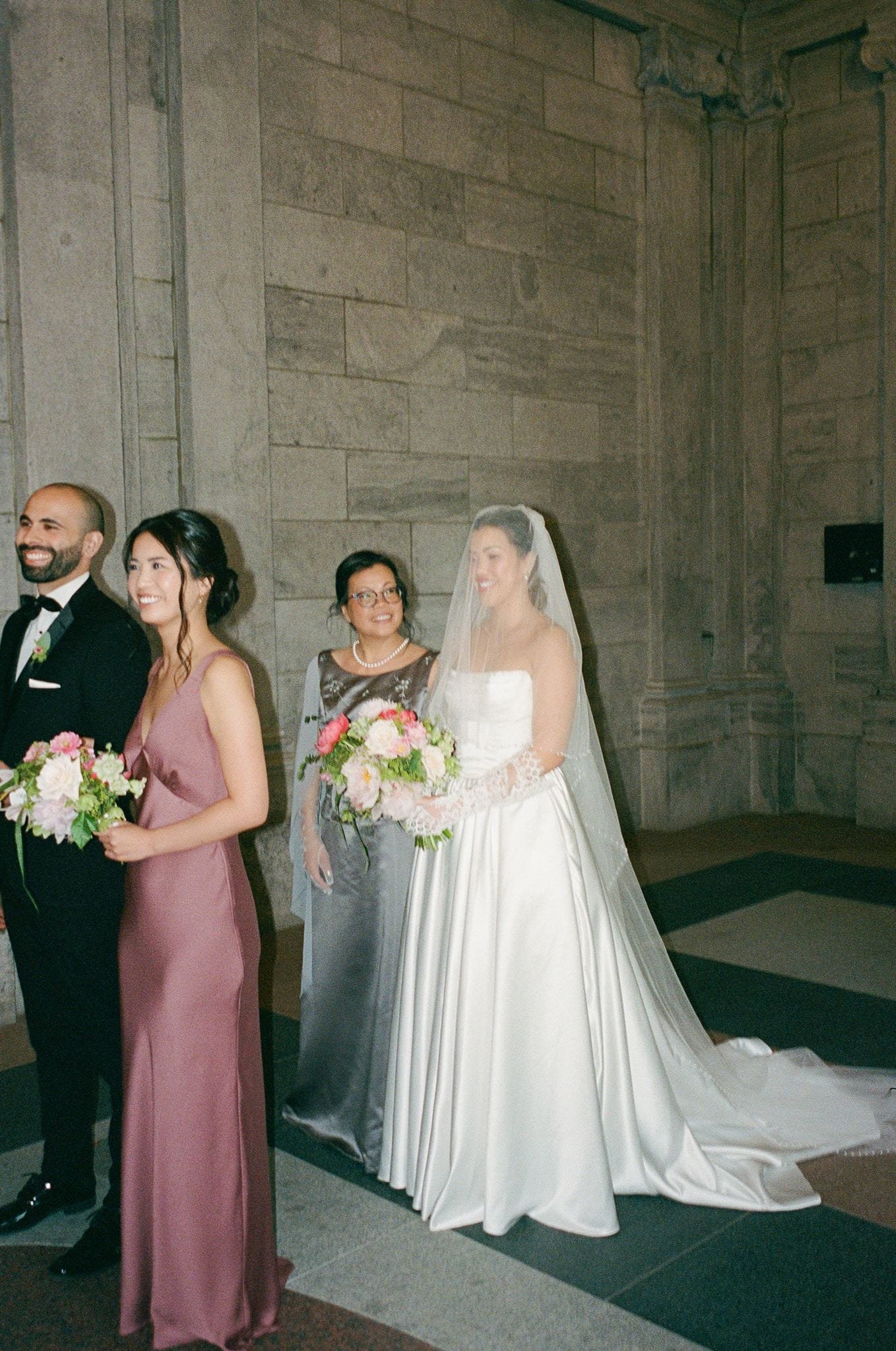 Group of people, including a bride in a white dress with veil, holding bouquet, and bridesmaid in mauve dress, smiling inside a church.