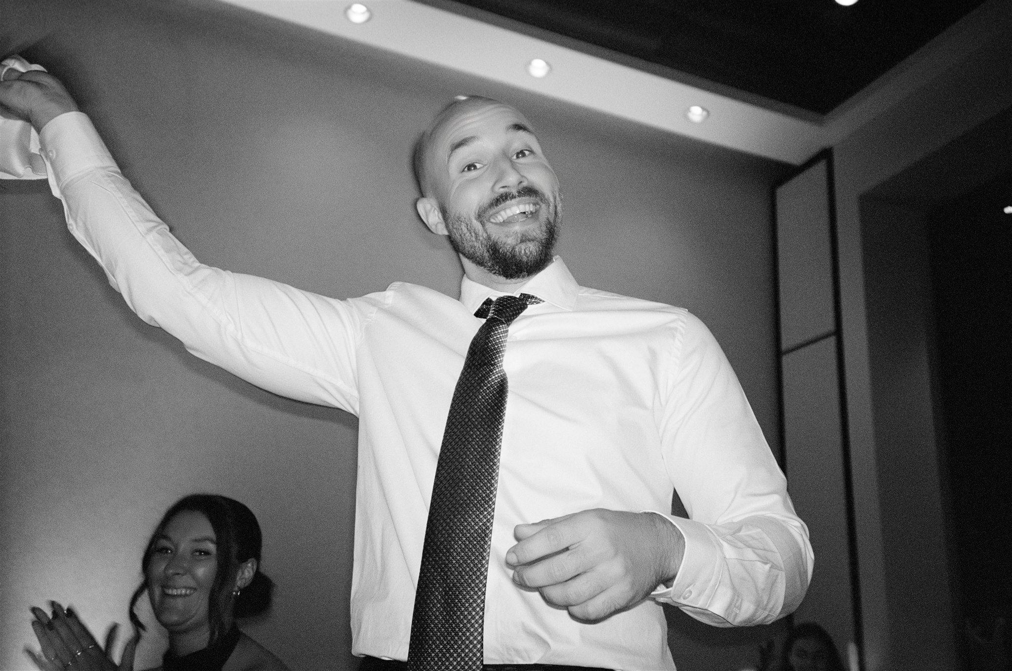 Homme souriant en costume blanc et cravate, levant le bras, avec une femme en arrière-plan applaudissant.