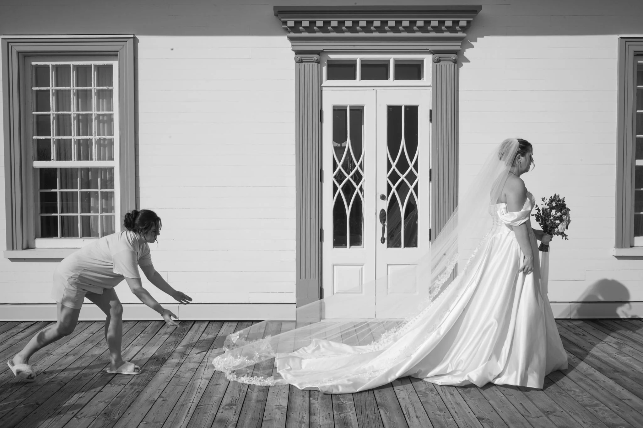 Une bride en robe de mariée avec un voile est assistée par une femme pliée en deux, qui semble l'aider ou ajuster sa robe, devant une façade de maison blanche avec fenêtres à volets.