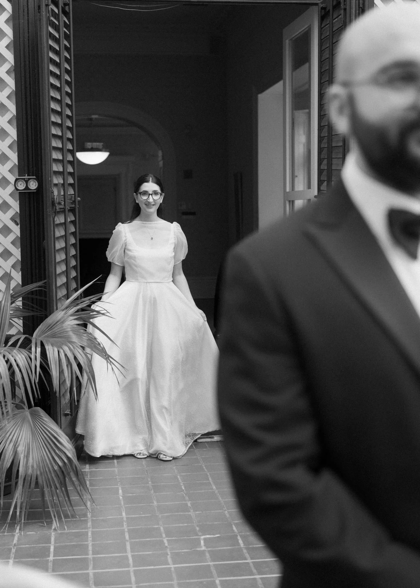 Une jeune femme portant une robe blanche et des lunettes sourit, vue à travers un reflet ou une fenêtre, en présence d'une personne floue en costume à l'avant-plan.