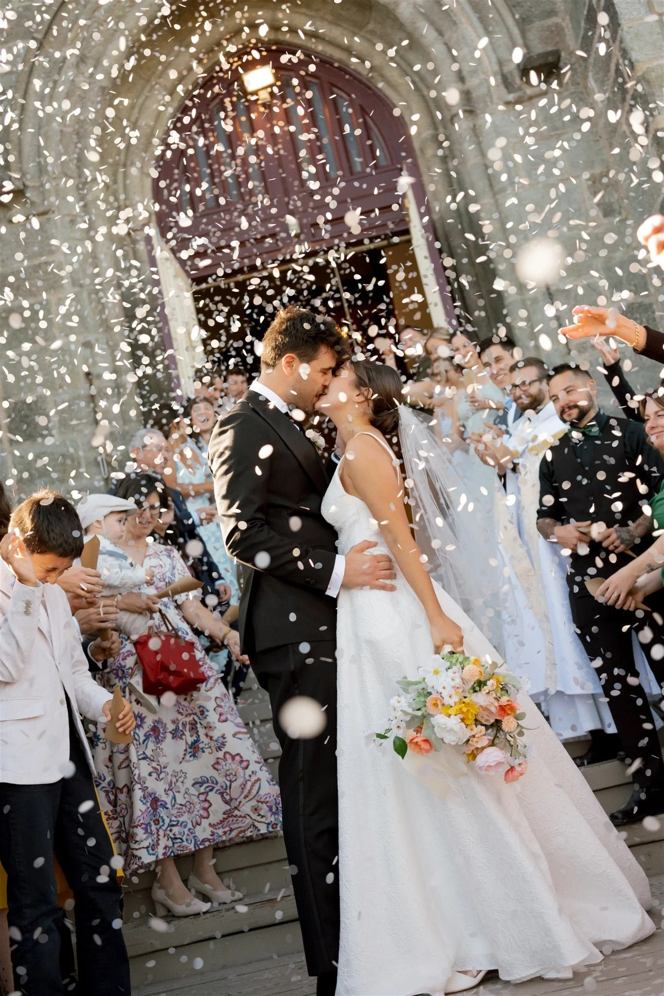Couple de mariés s’embrassant sous une pluie de confettis devant un bâtiment en pierre, entourés d’invités en pleine célébration, captés par un Photographe mariage Québec devant une église à Québec.
