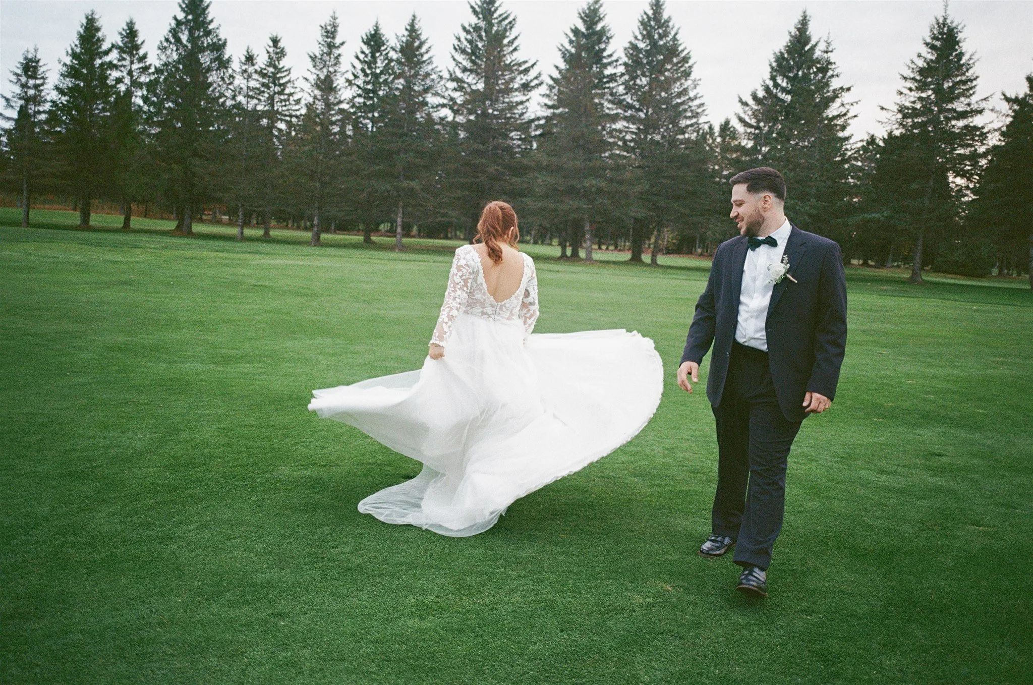 Un couple lors d'un mariage en plein air, la femme en robe de mariée blanche danse ou marche sur l'herbe, tandis que l'homme en costume noir avec une cravate noire la regarde avec un sourire, entouré de grands arbres dans un parc ou une forêt.