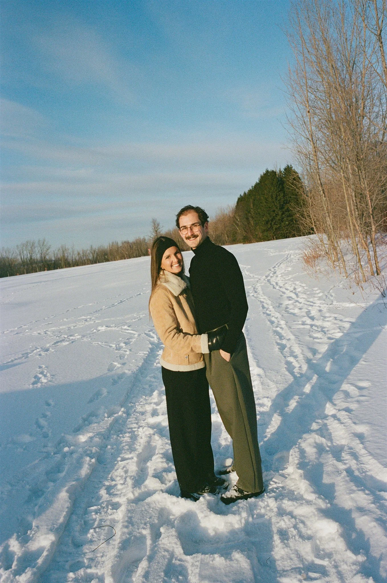 Un couple souriant dans un paysage enneigé avec des arbres nus et un ciel bleu clair.