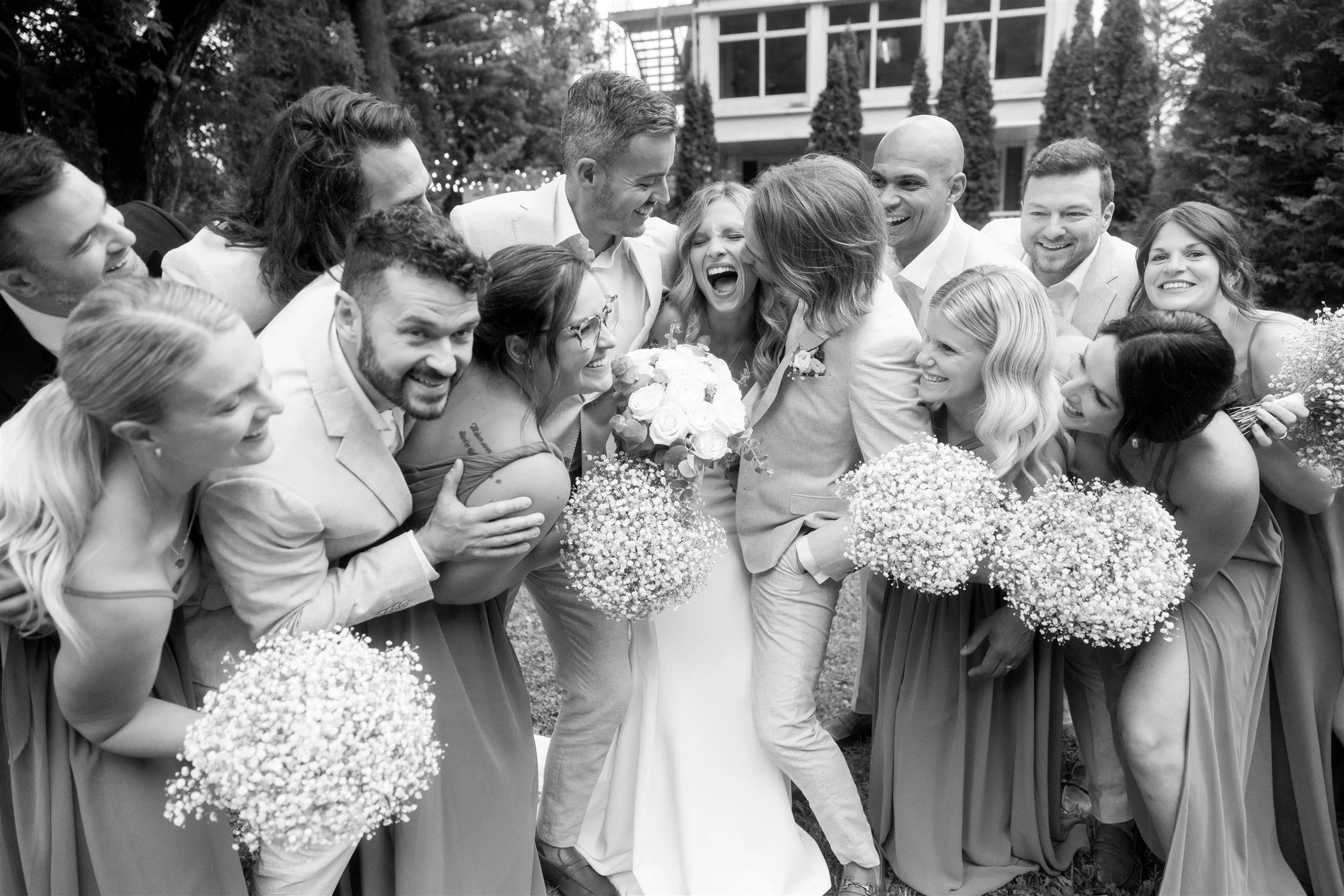 Groupe de personnes lors d'un mariage, souriantes et en train de célébrer dehors, avec des robes de demoiselles d'honneur, bouquets de fleurs, et un couple de mariés au centre.