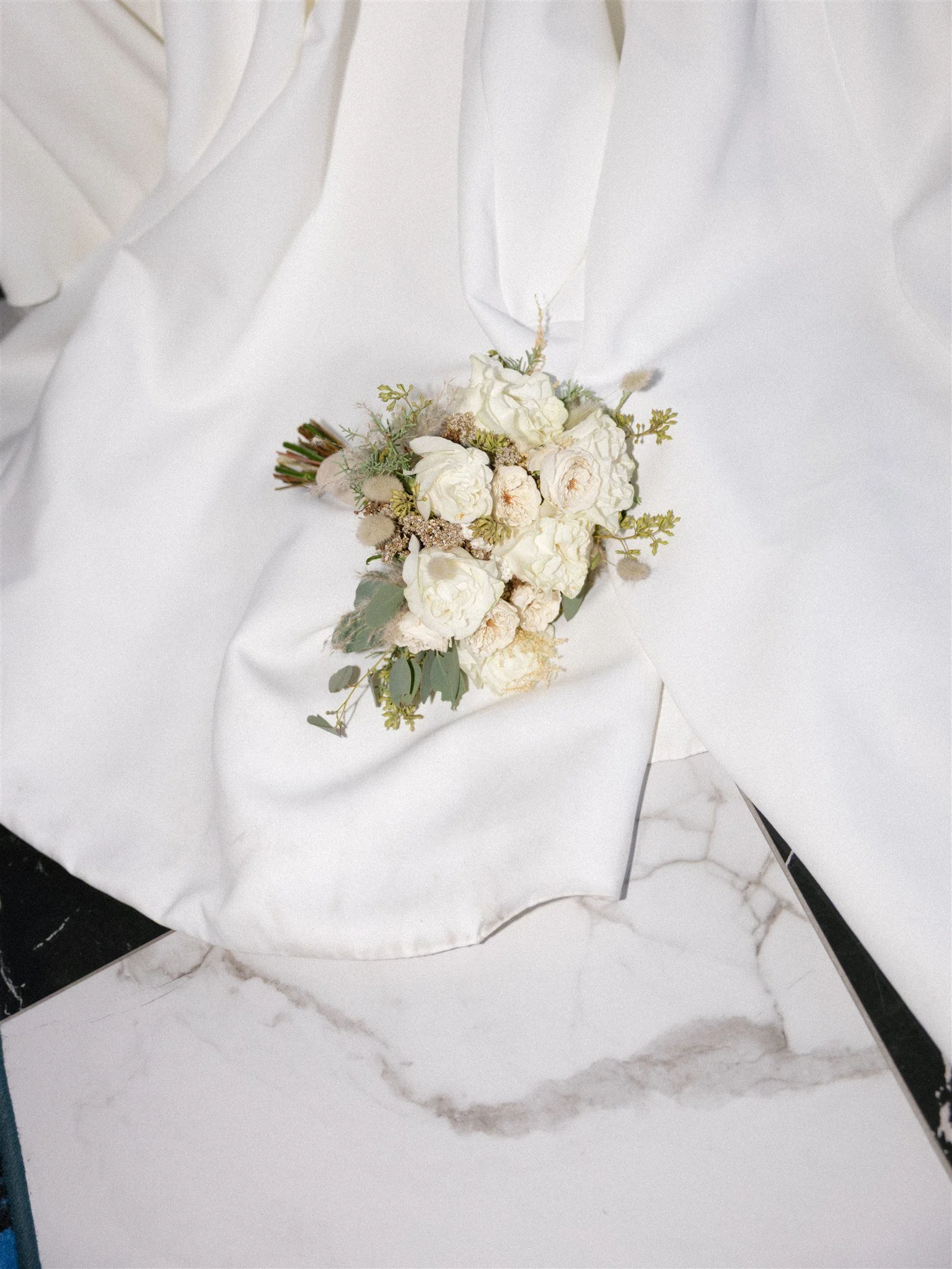 Bouquet de fleurs blanches posé sur un drap blanc en tissu satiné, avec un sol en marbre blanc et noir en arrière-plan, capté par un Photographe mariage Québec.