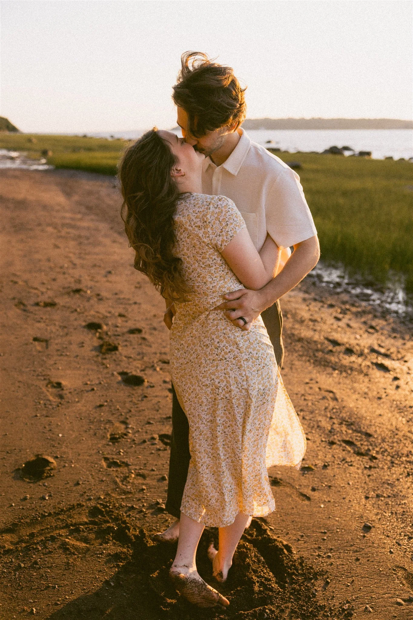 Un couple s'embrasse tendrement sur une plage au coucher du soleil, en tenues décontractées, avec la mer et le ciel lumineux en arrière-plan.